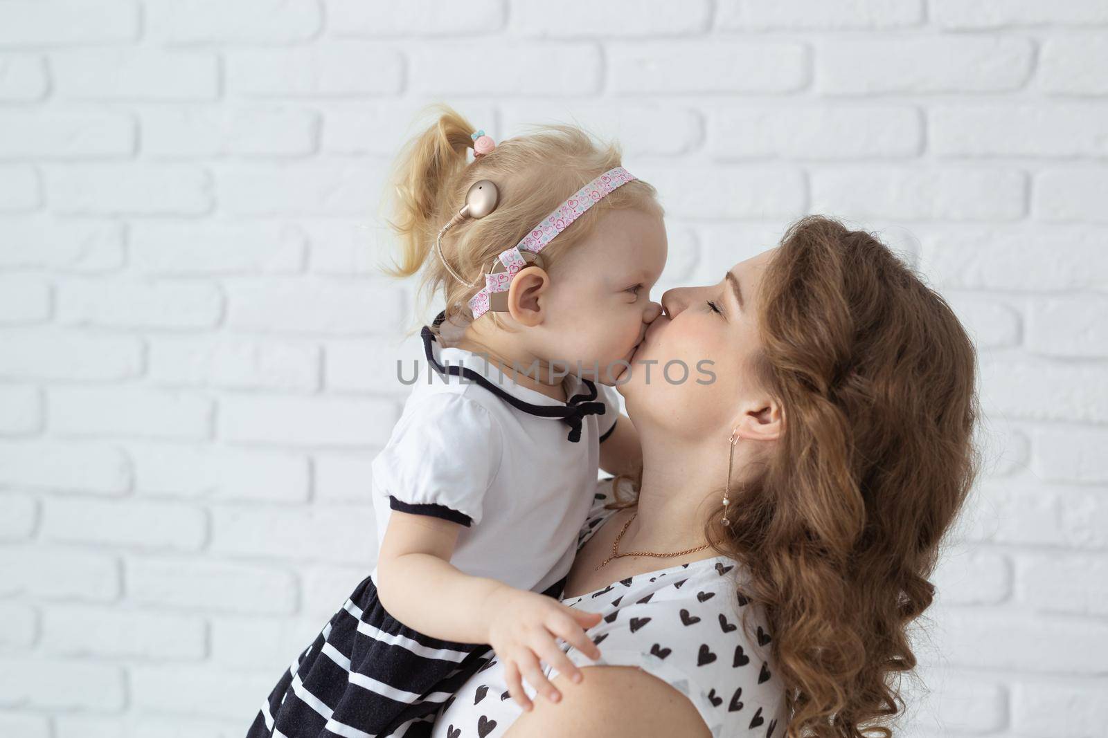 Mother holds and having fun child with hearing aids and cochlear implants on white brick wall with copy space . Deaf and diversity and innovating medicine technologies concept by Satura86