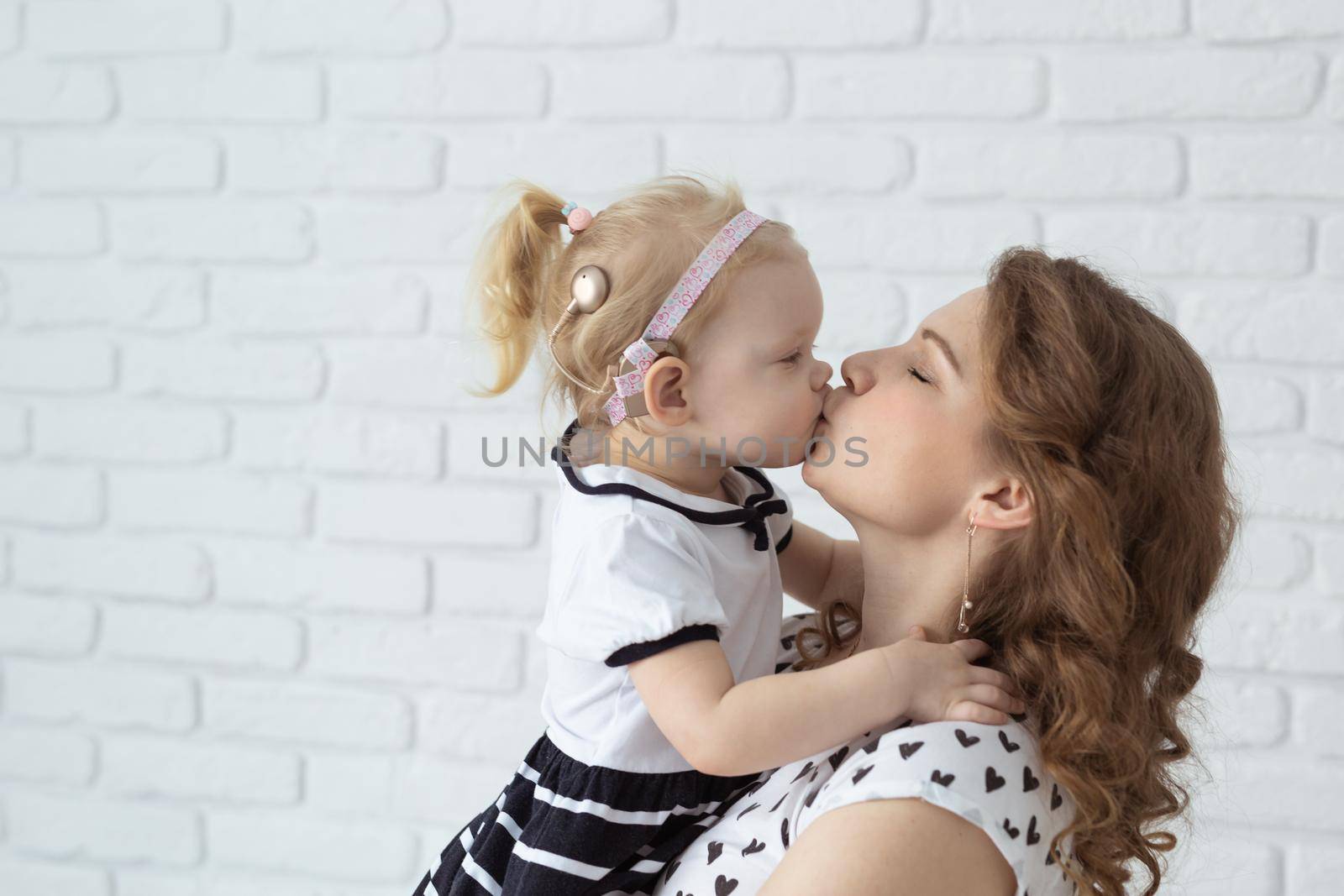 Mother holds and having fun her child with hearing aids and cochlear implants on white brick wall with copy space and place for advertising . Deaf and innovating medicine technologies concept by Satura86