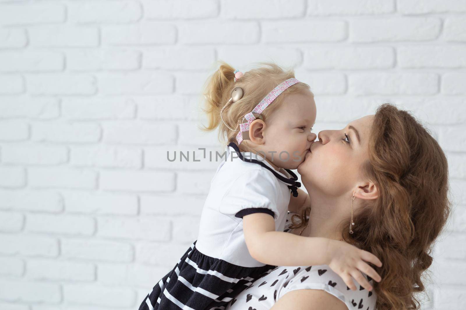 Mother holds and having fun child with hearing aids and cochlear implants on white brick wall with copy space . Deaf and diversity and innovating medicine technologies concept by Satura86