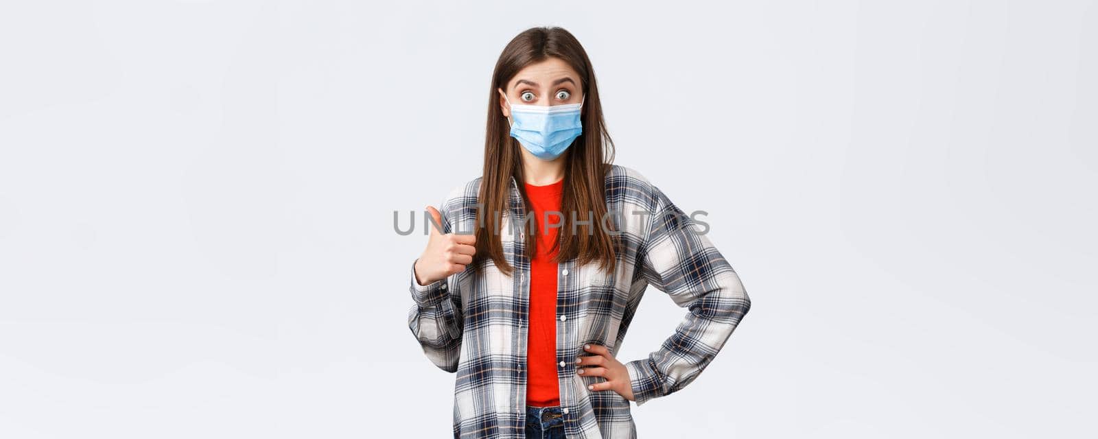 Coronavirus outbreak, leisure on quarantine, social distancing and emotions concept. Excited and surprised young woman in medical mask hear really good idea, show thumb-up in approval.