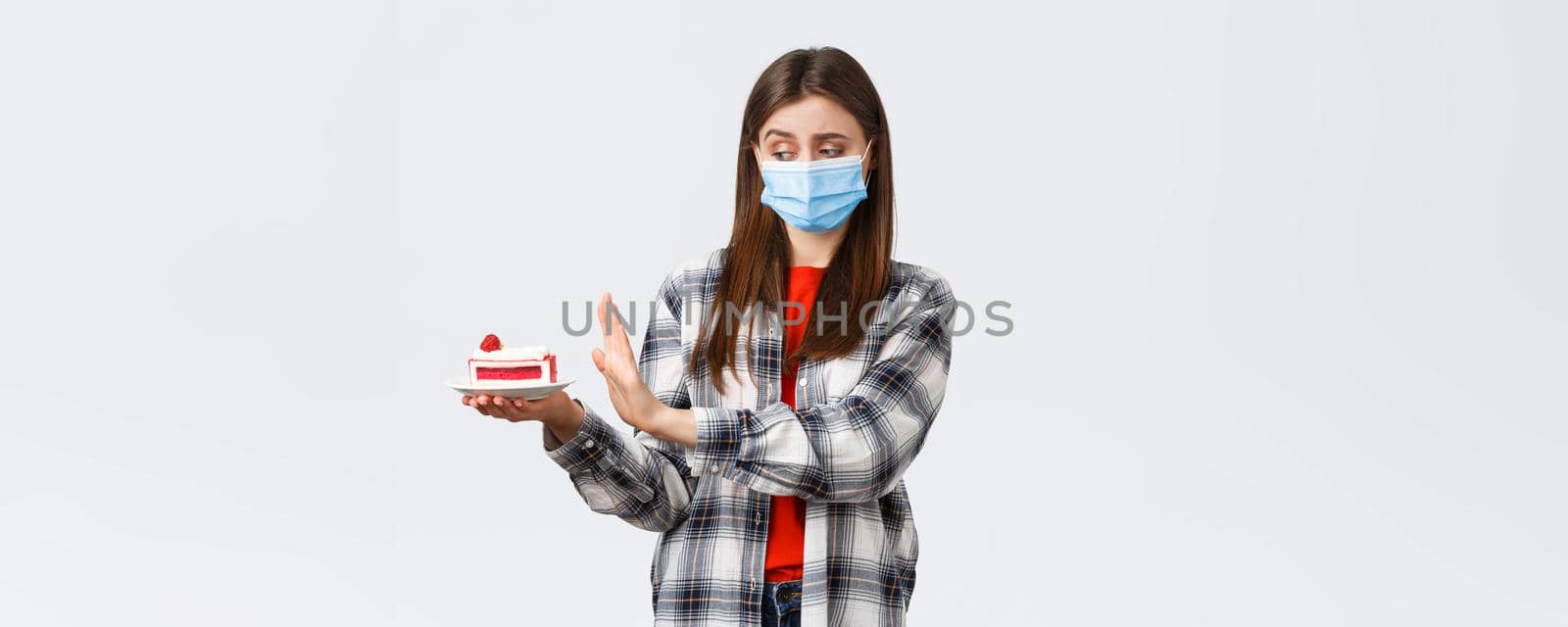 Coronavirus outbreak, lifestyle during social distancing and holidays celebration concept. Displeased and reluctant cute girl in medical mask refuse eat cake, show stop sign at dessert.