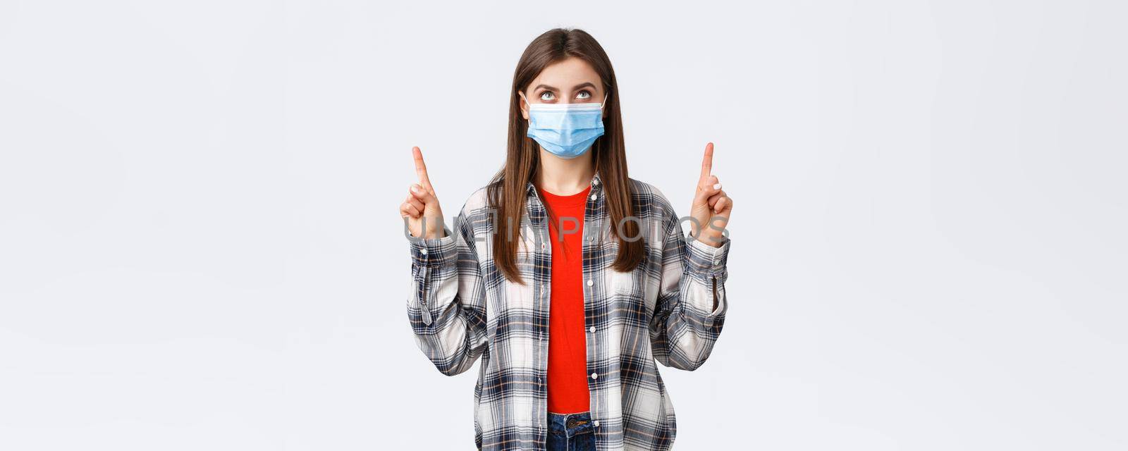 Coronavirus outbreak, leisure on quarantine, social distancing and emotions concept. Interested serious young woman in medical mask and casual outfit, looking and pointing up banner.