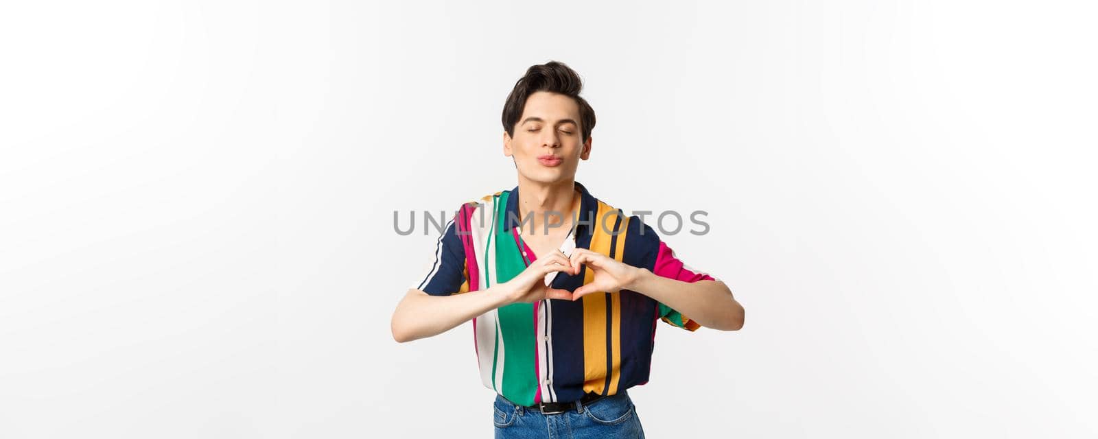 Lovely young man pucker lips and showing heart sign, waiting for kiss, I love you gesture, standing over white background.