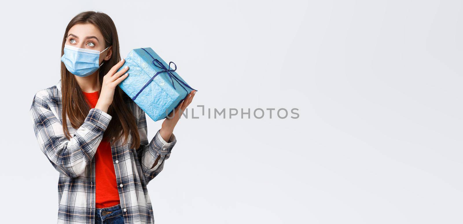 Covid-19, lifestyle, holidays and celebration concept. Curious young cute girl celebrating birthday, shaking gift box to guess what inside, look away thoughtful, lean ear to present, white background.