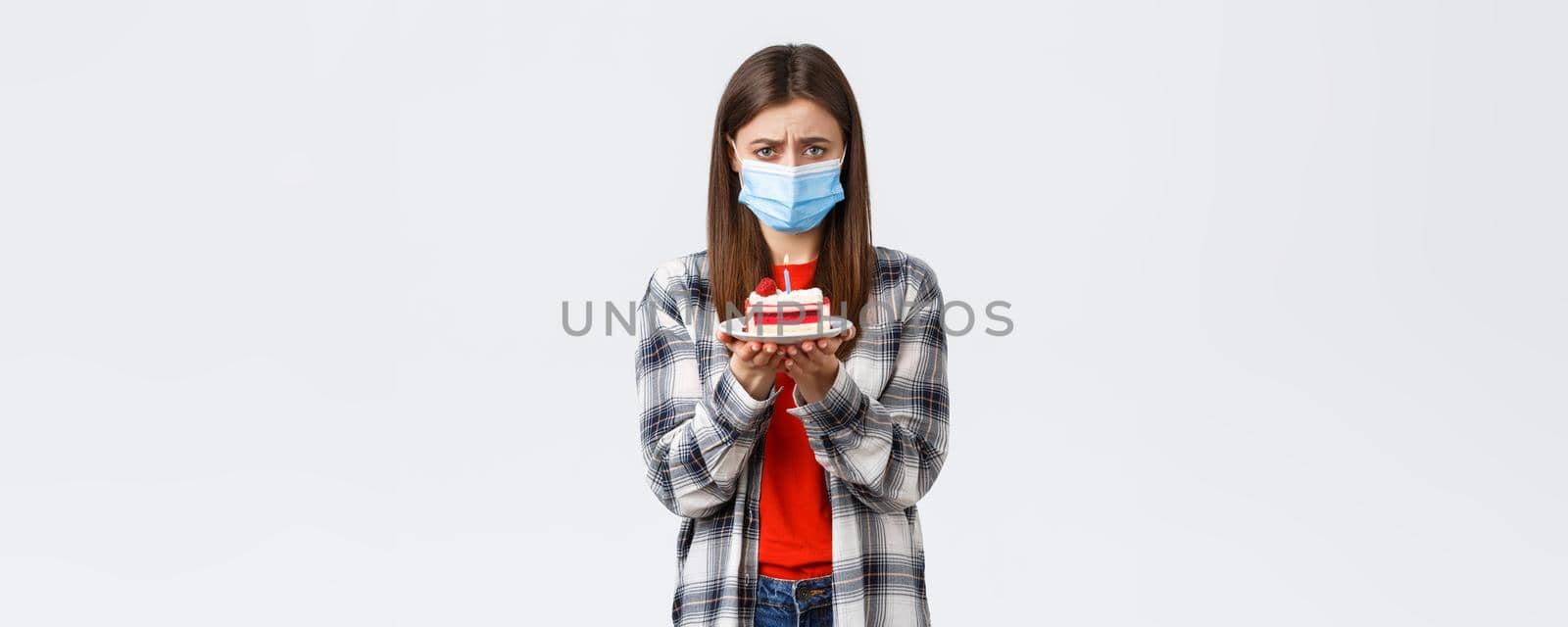 Coronavirus outbreak, lifestyle on social distancing and holidays celebration concept. Lonely and upset cute girl in medical mask, celebrating birthday all alone on self-quarantine, sob and hold cake by Benzoix