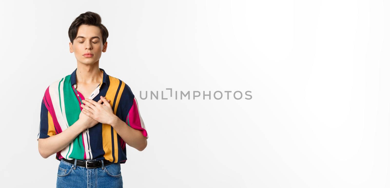 Image of nostalgic young gay man standing with closed eyes and hands on heart, remember something, standing over white background.