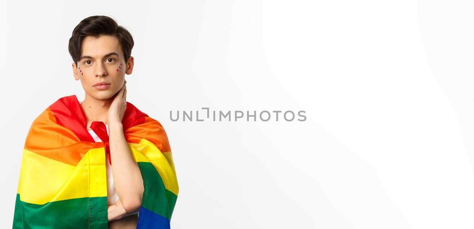 Waist-up shot of beautiful gay man with glitter on face, wrap himself with rainbow lgbt flag and touching face gently, looking at camera, white background.