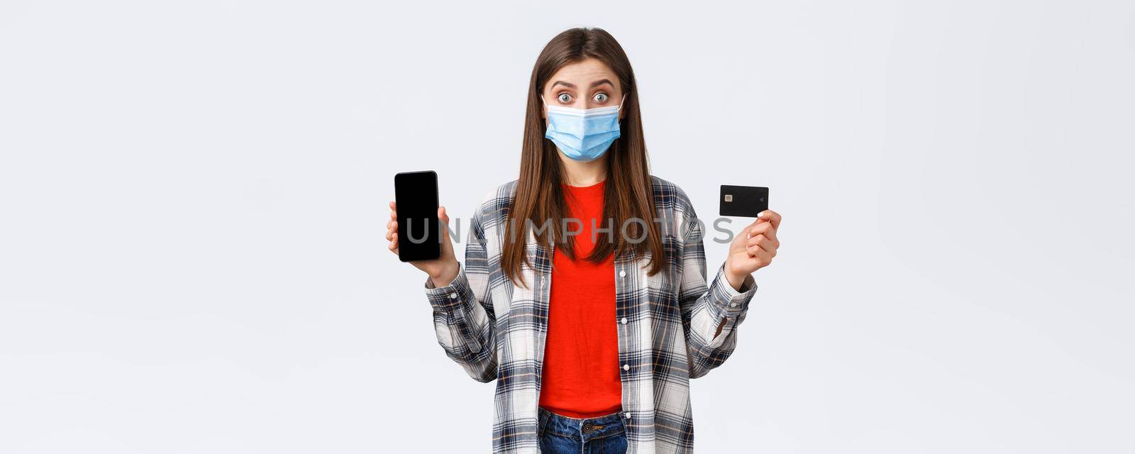 Coronavirus outbreak, working from home, online shopping and contactless payment concept. Excited woman in medical mask showing credit card and mobile phone, look surprised by Benzoix