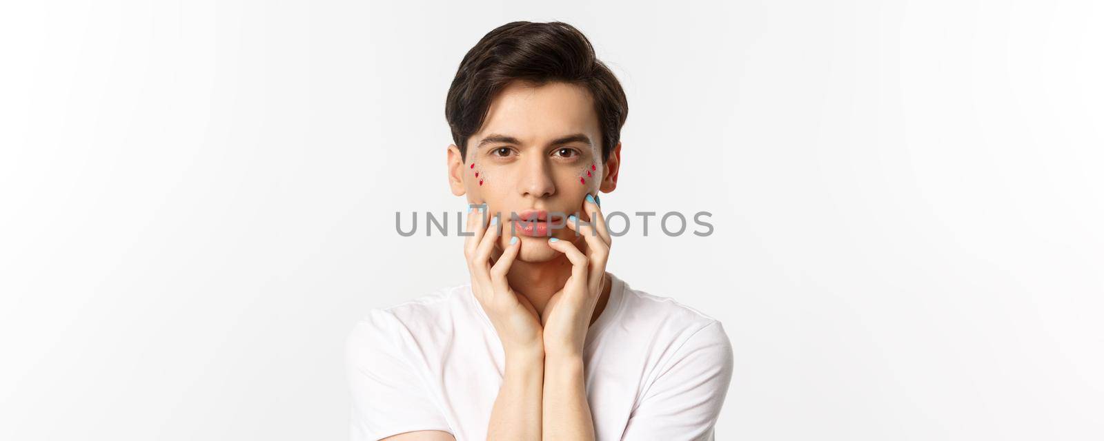 People, lgbtq and beauty concept. Close-up of beautiful queer man touching face with fingers with blue nail polish, standing over white background by Benzoix