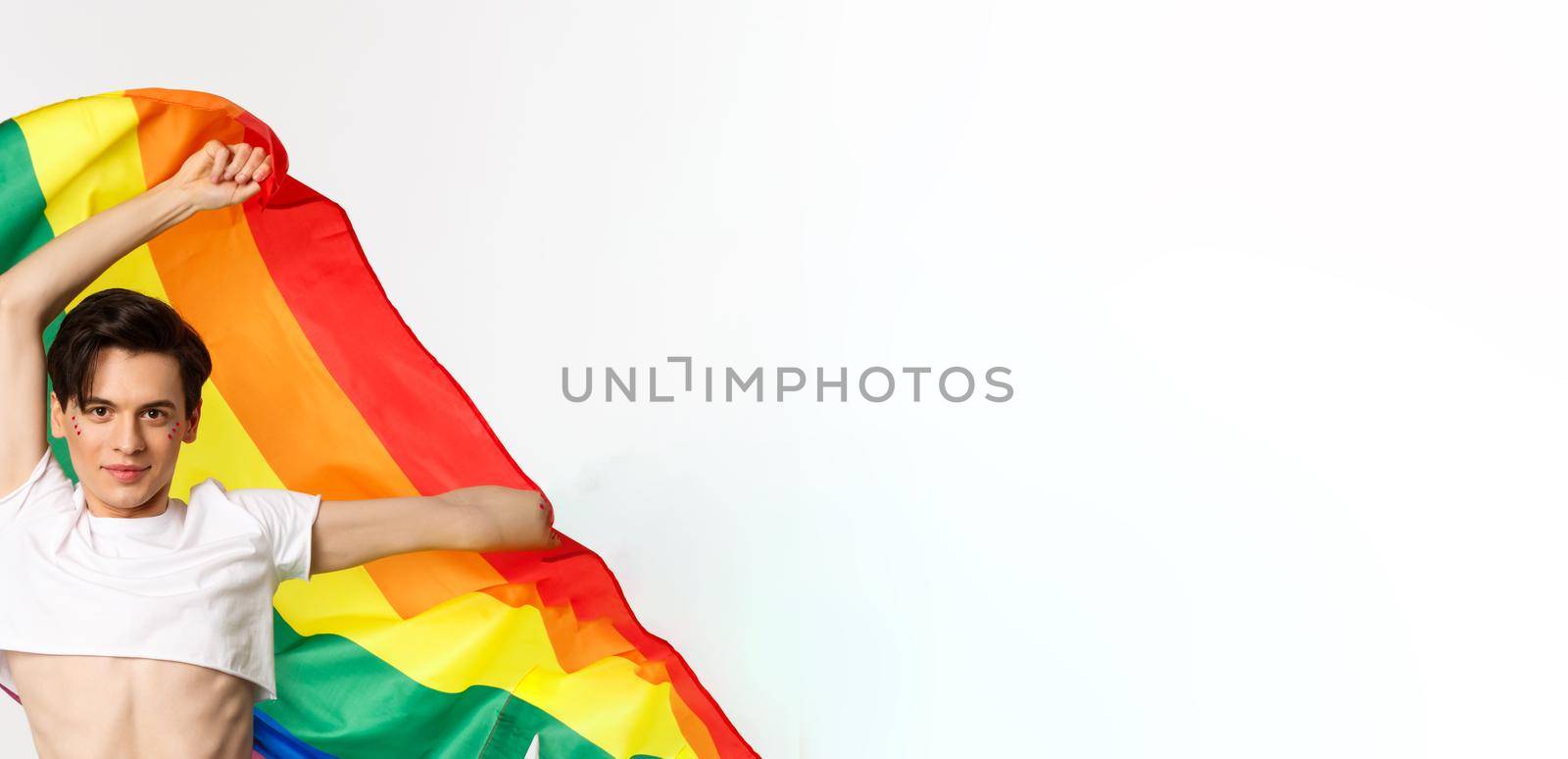 Vertical view of happy queer person in crop top and jeans waving raised rainbow flag, celebrating lgbtq holiday, standing over white background.