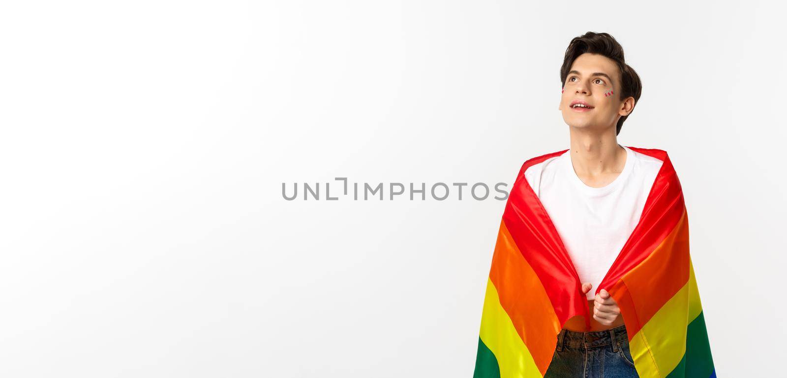 Human rights and lgbtq community concept. Beautiful young androgynous man with glitter on face, wearing rainbow pride flag and looking at upper left corner, white background.