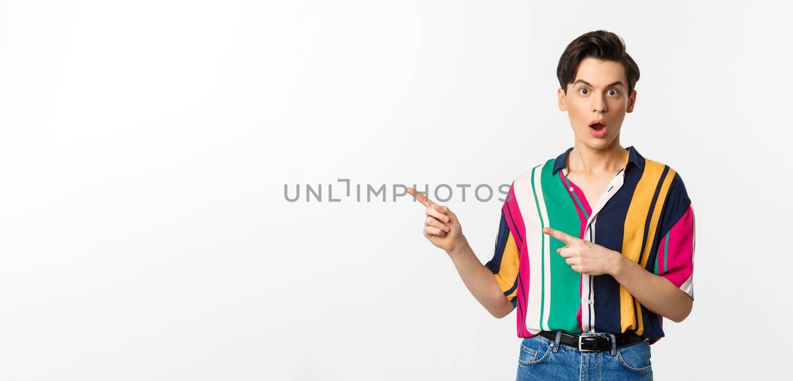 Amazed young queer man pointing fingers left, staring at camera with disbelief, asking question about product, standing over white background by Benzoix