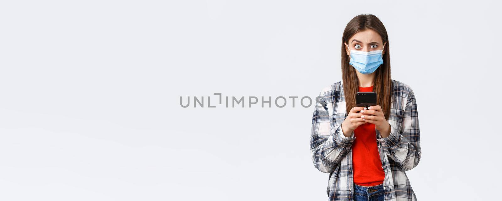 Different emotions, covid-19, social distancing and technology concept. Puzzled and confused young woman in medical mask react to strange message, hold mobile phone, look camera unsure.