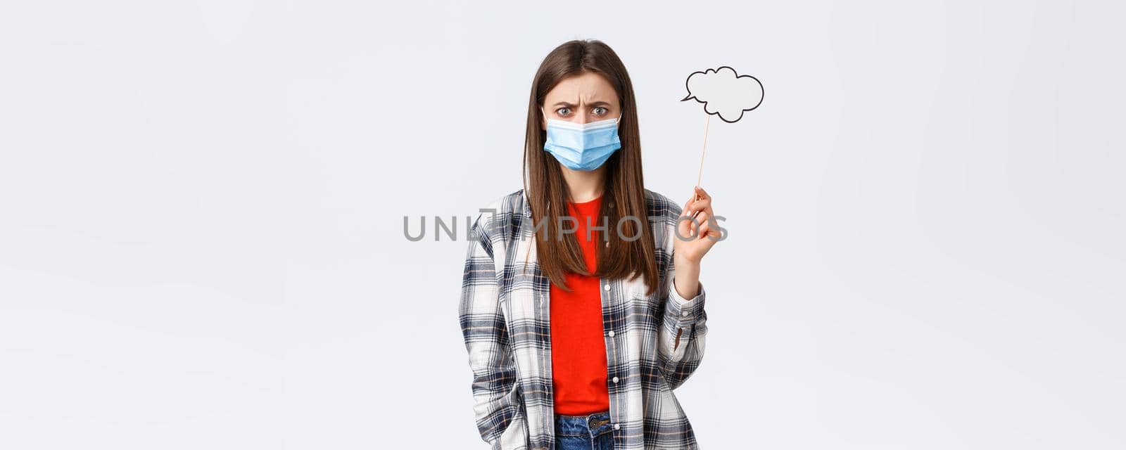 Coronavirus outbreak, leisure on quarantine, social distancing and emotions concept. Troubled young woman in medical mask frowing upset or disappointed, hold comment cloud stick near head.
