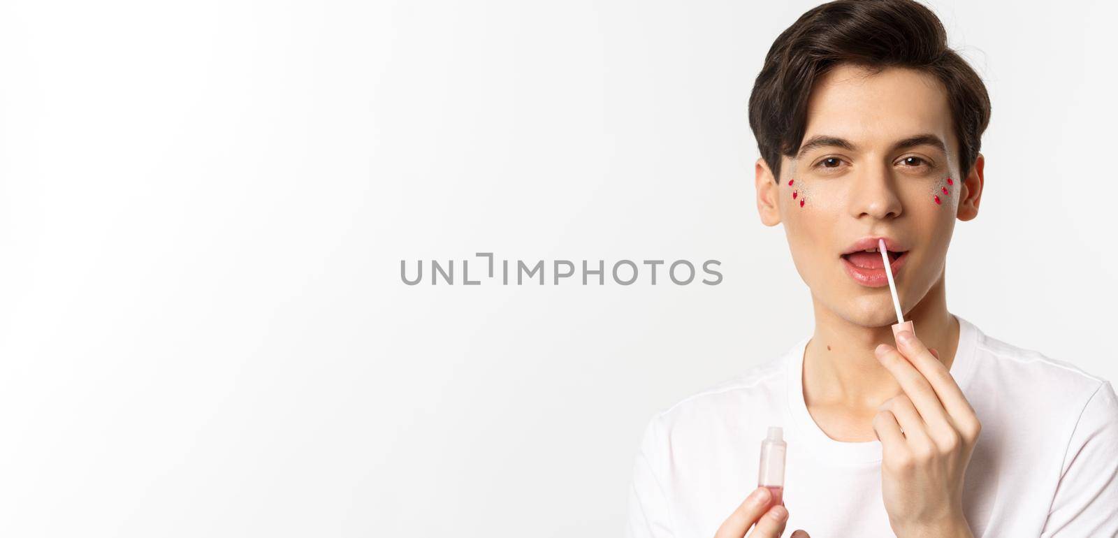 Close-up of gender fluid person with glitter on face, applying lip gloss lipstick and looking at camera, standing against white background by Benzoix