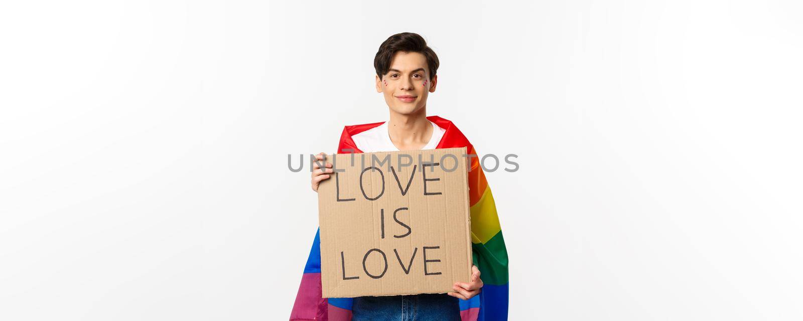 Smiling gay man activist holding sign love is love for lgbt pride parade, wearing Rainbow flag, standing over white background. Copy space