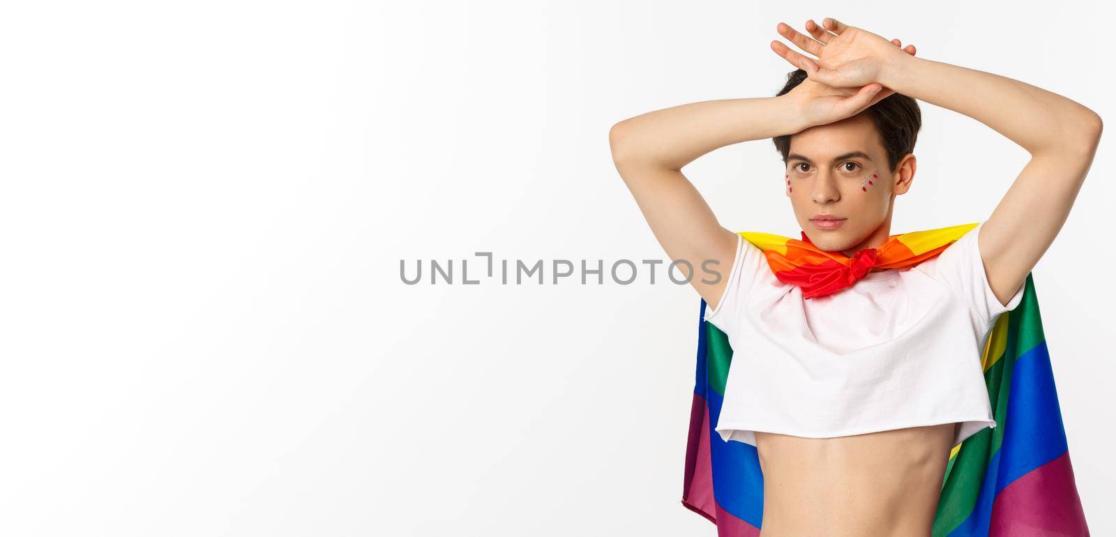 Beautiful gay man with glitter on face, wearing crop top and rainbow lgbt flag, posing against white background by Benzoix