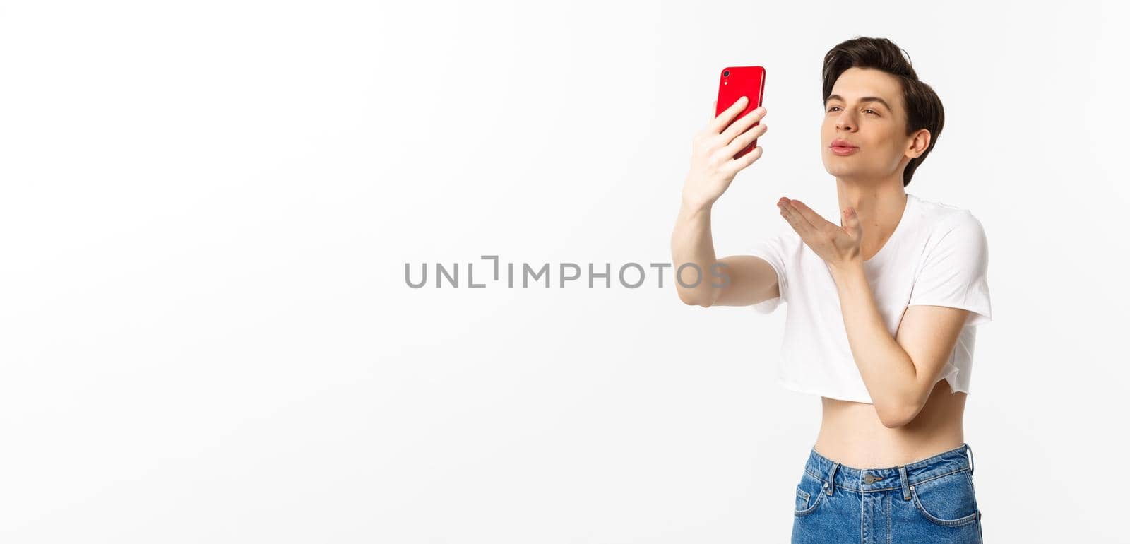 Beautiful gay man in crop top sending air kiss at phone camera, taking selfie or video chat on smartphone, standing over white background.
