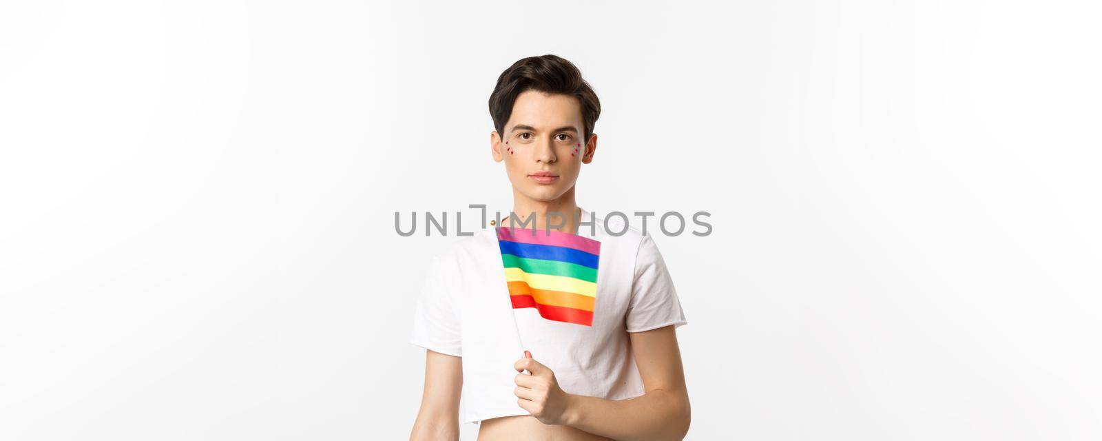 Pride and lgbtq concept. Waist up shot of attractive anrogynous man holding rainbow flag, having glitter on face and looking at camera, standing over white background.