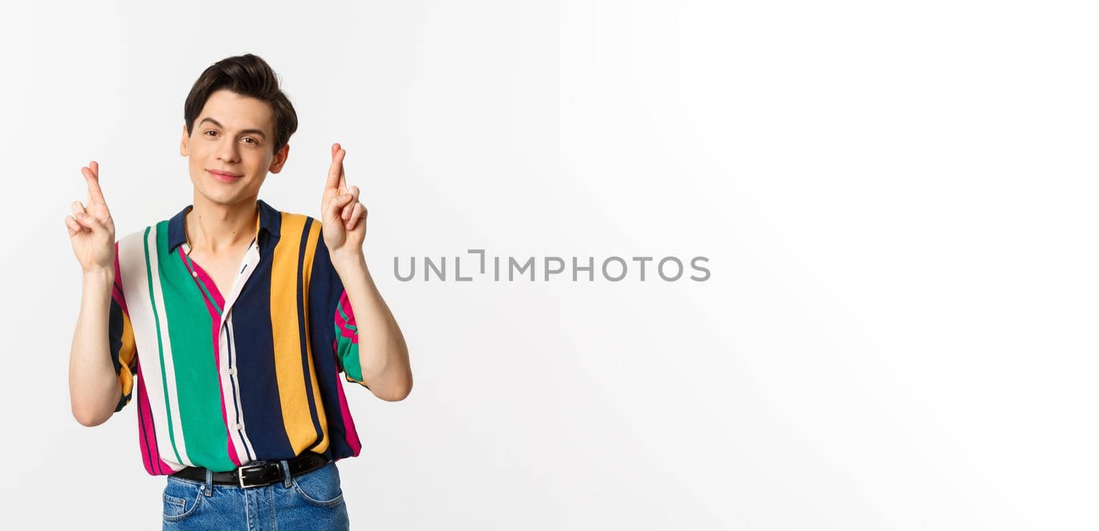 Optimistic young man smiling, holding fingers crossed for good luck, making wish, standing over white background. Copy space