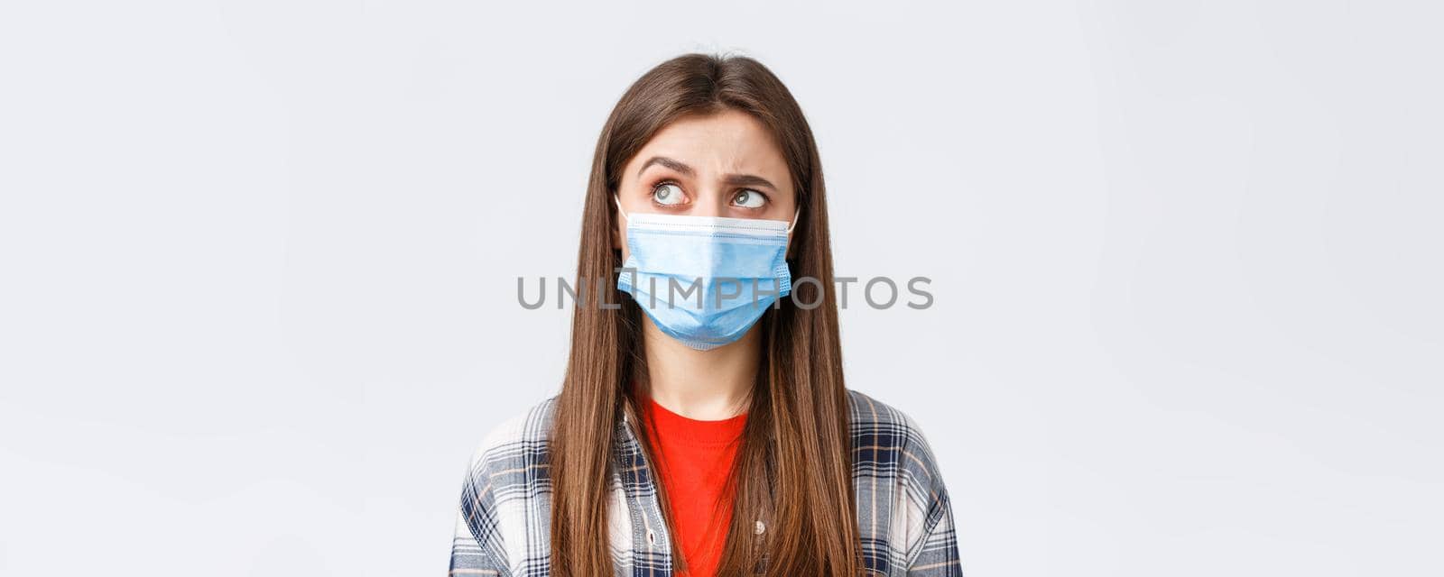 Coronavirus outbreak, leisure on quarantine, social distancing and emotions concept. Intrigued and hesitant young woman in medical mask, looking upper left corner reluctant, white background by Benzoix