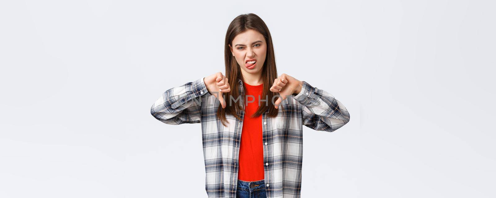 Lifestyle, different emotions, leisure activities concept. Displeased and unimpressed young picky girl in checked shirt, thumb-down and show tongue to express dislike, rate awful movie by Benzoix