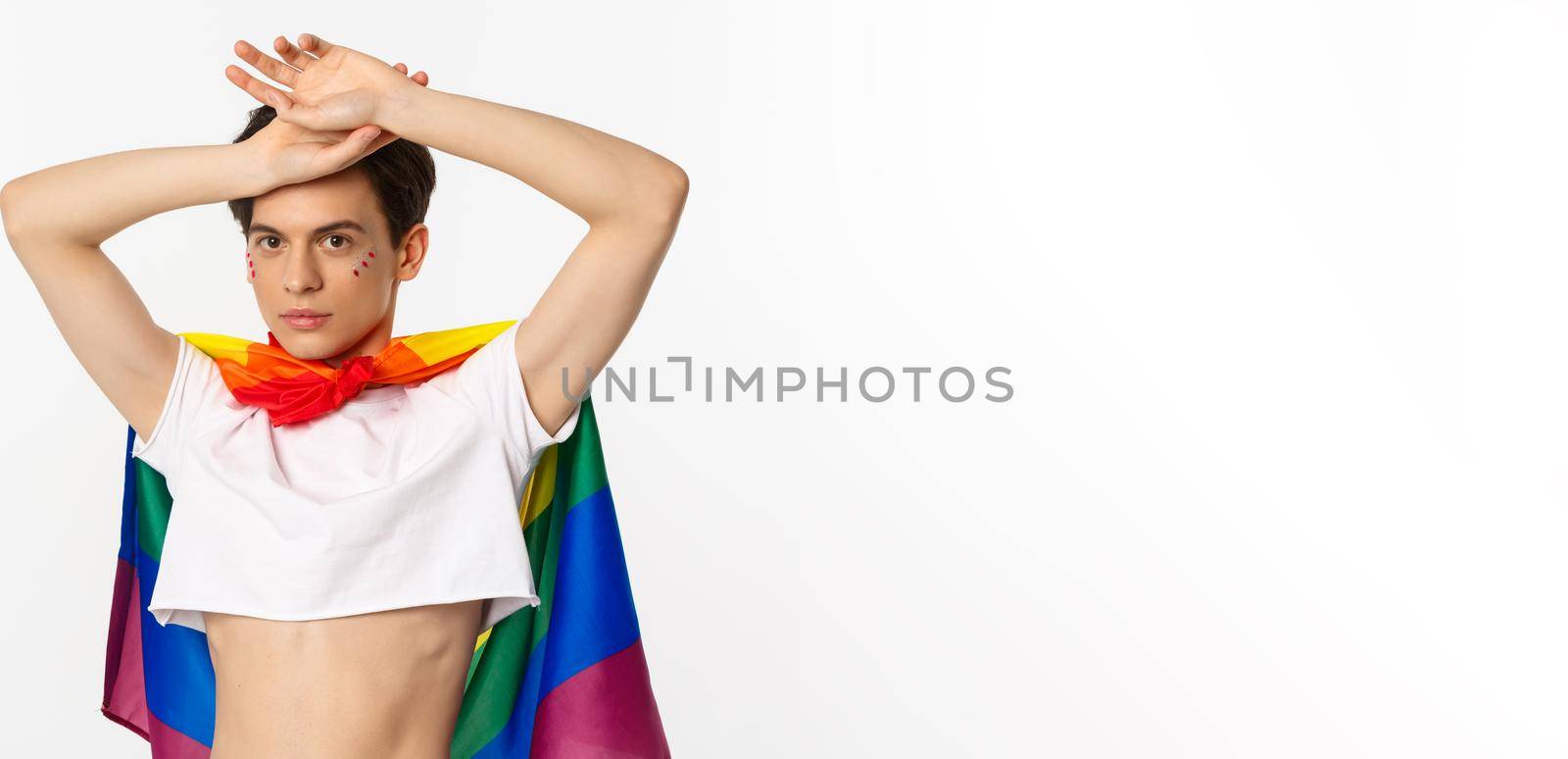 Beautiful gay man with glitter on face, wearing crop top and rainbow lgbt flag, posing against white background.