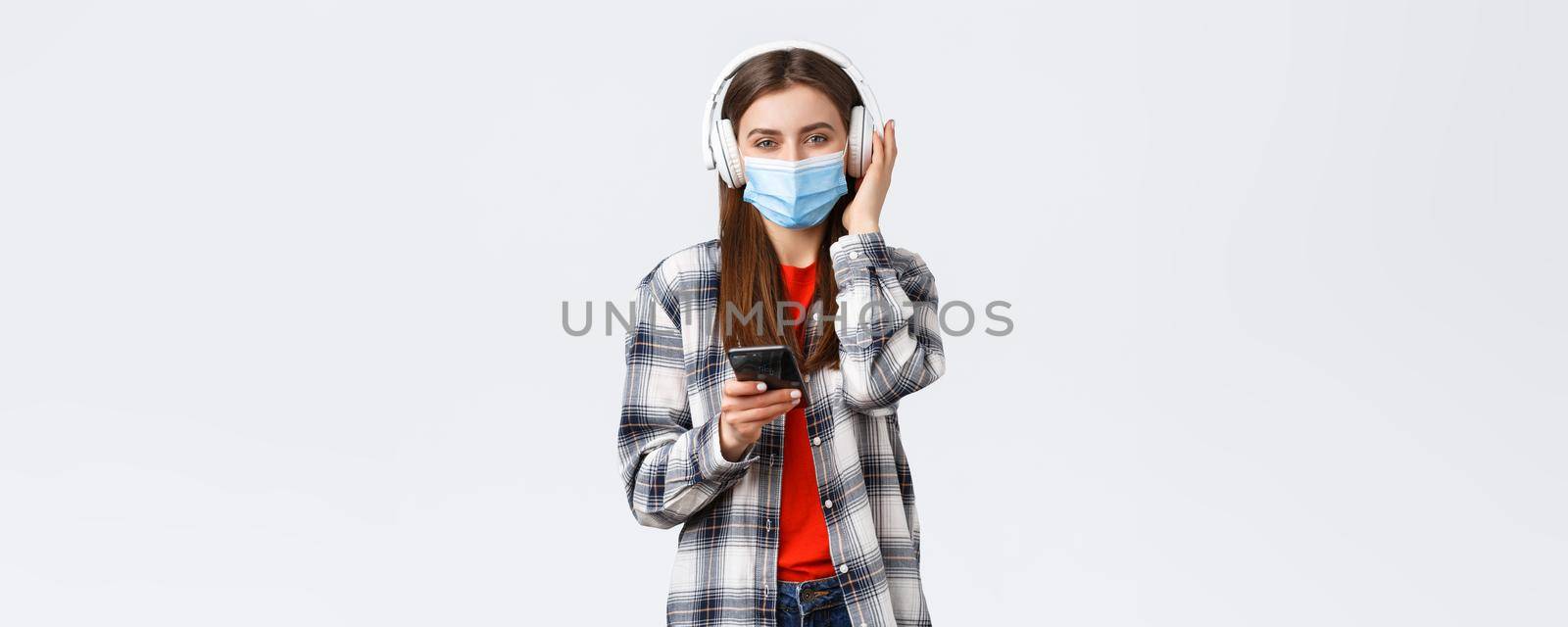 Social distancing, leisure and lifestyle on covid-19 outbreak, coronavirus concept. Cheerful smiling woman in medical mask, picking song from playlist smartphone, listen music in wireless headphones by Benzoix