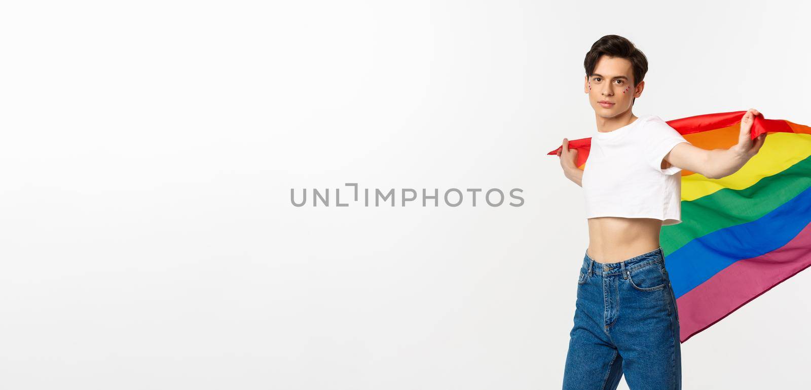 Human rights and lgbtq community concept. Out and proud gay man waving rainbow flag and looking confident at camera, standing in crop top and jeans against white background.