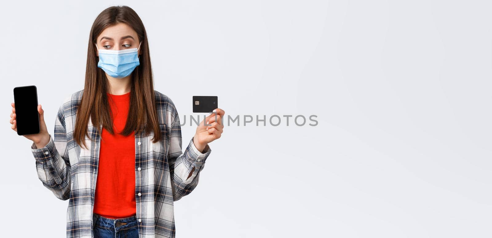 Coronavirus outbreak, working from home, online shopping and contactless payment concept. Girl in medical mask showing mobile phone and credit card, peek at screen making order by Benzoix