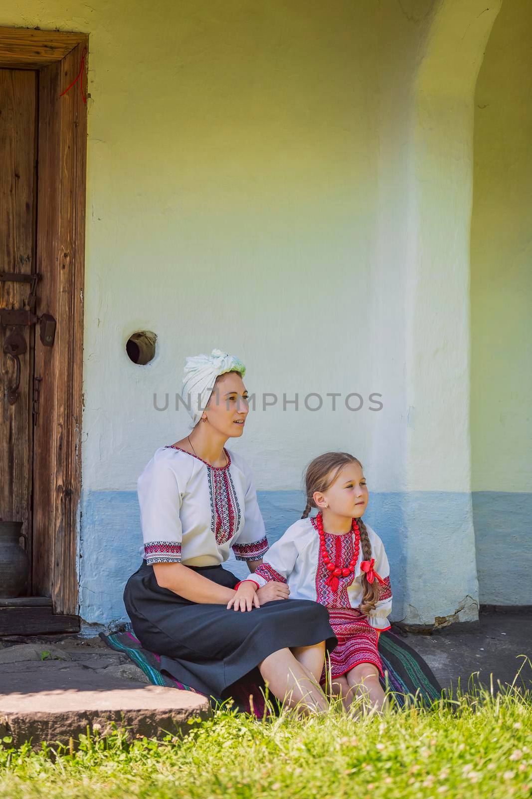 mother and daughter in Ukrainian national costumes are sitting near an old house