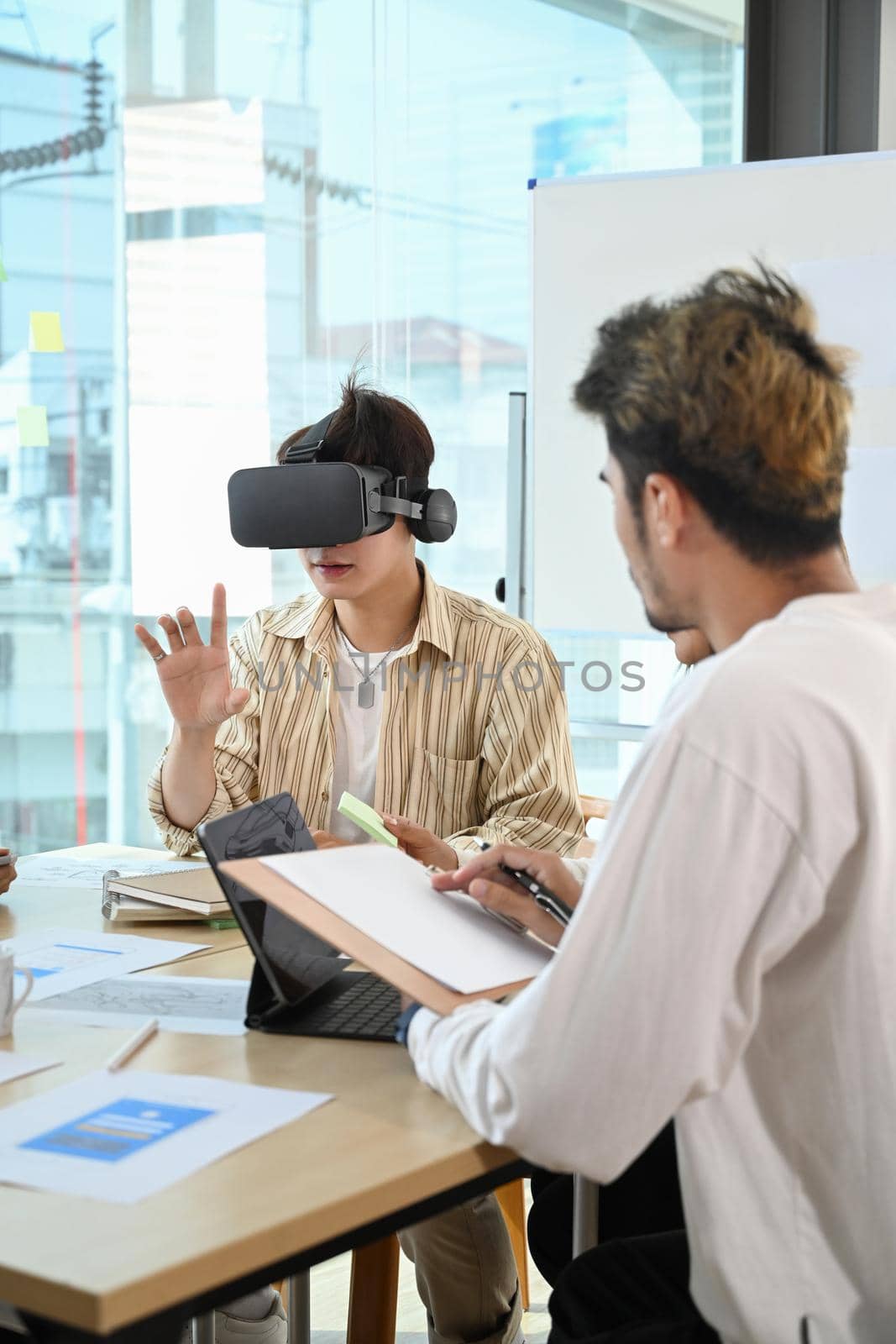 Young creative man wearing virtual reality headset and touching something while his colleagues discussing corporate project in meeting by prathanchorruangsak