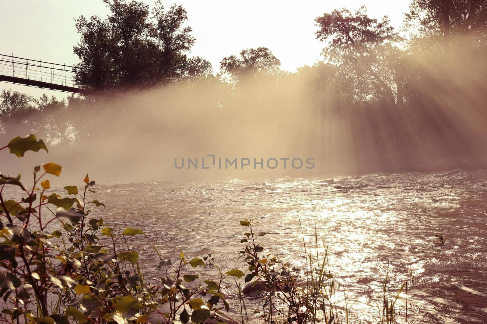 Foggy morning haze over the river with streaks of sunbeams, suspension footbridge, morning. Toned image by Proxima13