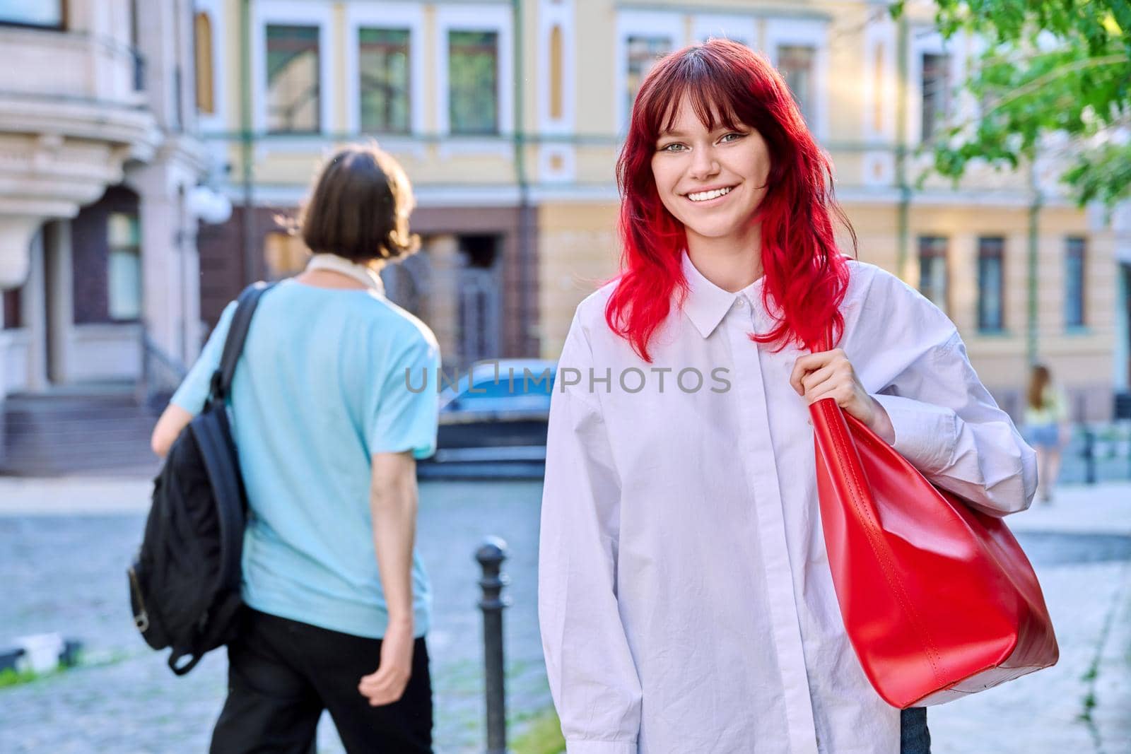 Teenage fashionable smiling female with red dyed hair looking at camera, urban style, street background, copy space. Youth, fashion, beauty, trends, people concept