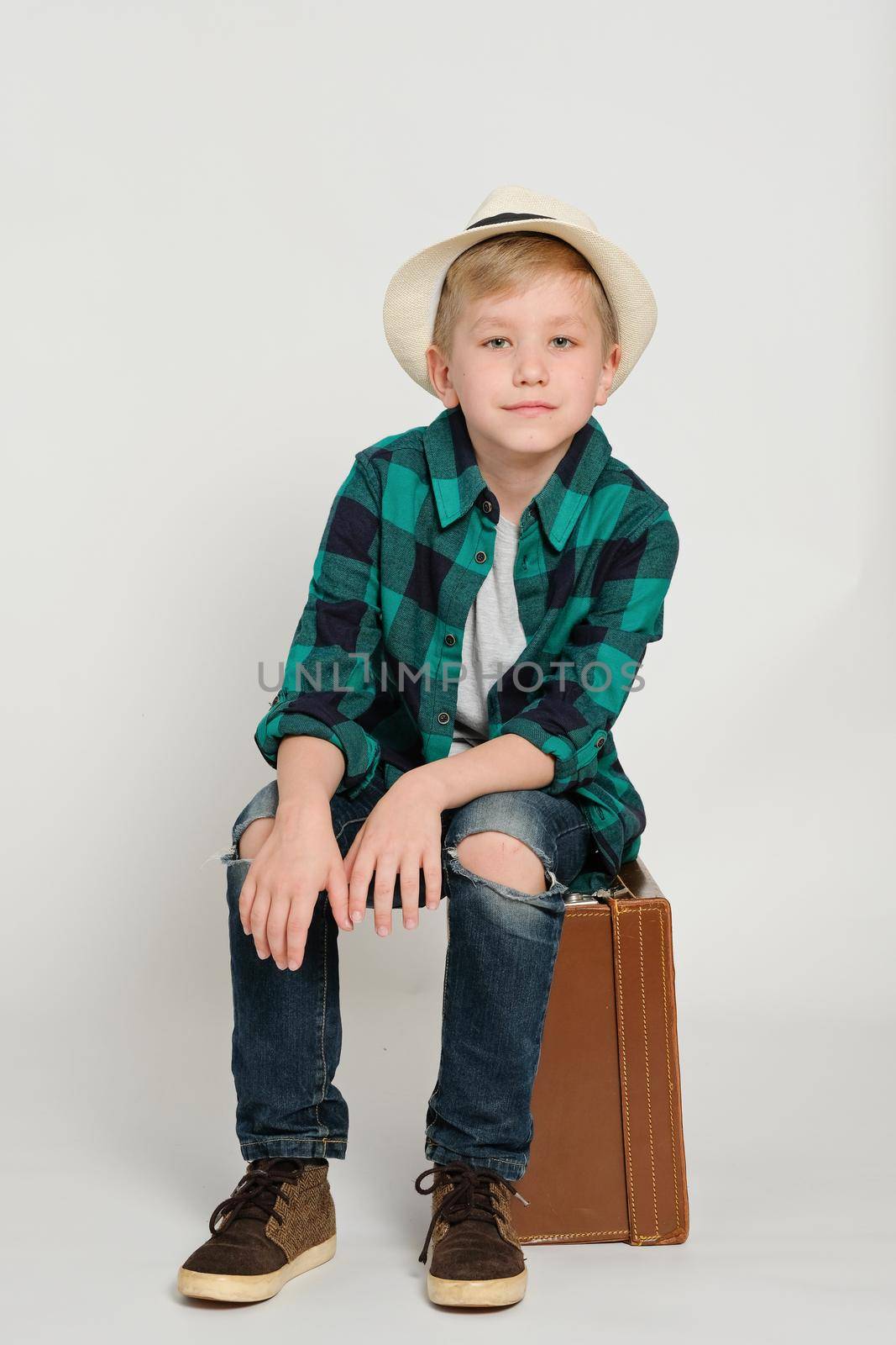 Portrait of cute stylish blond boy kid 7 years old wearing hat and checked shirt and jeans sitting on a retro suitcase. Travel concept by natus111