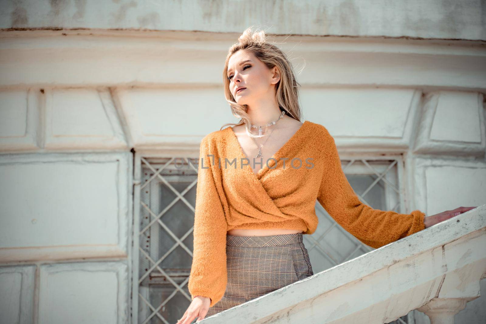 A middle-aged woman looks like a good blonde with curly beautiful hair and makeup on the background of the building. She is wearing a yellow sweater