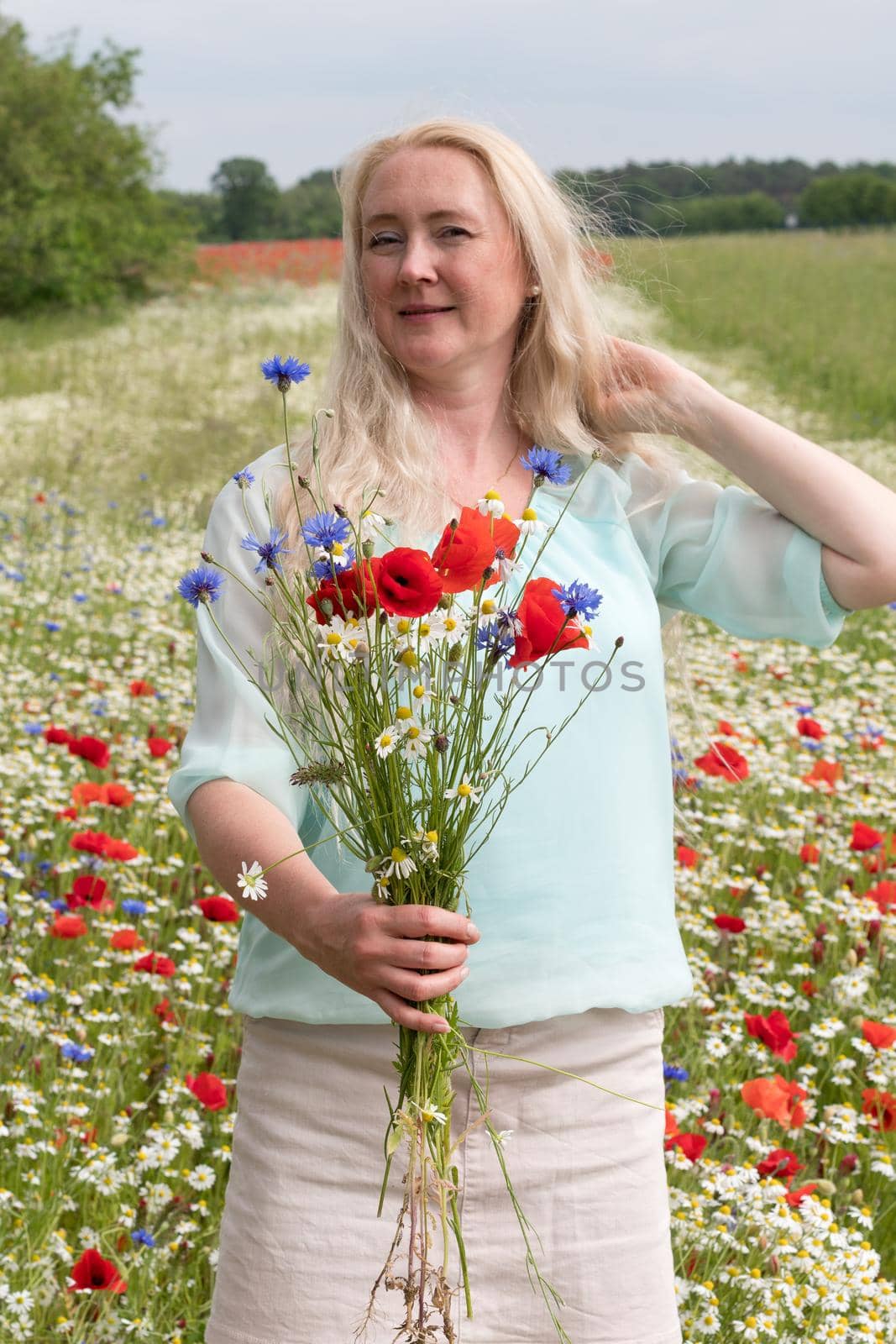 beautiful middle-aged blonde woman stands among a flowering field of poppies by KaterinaDalemans