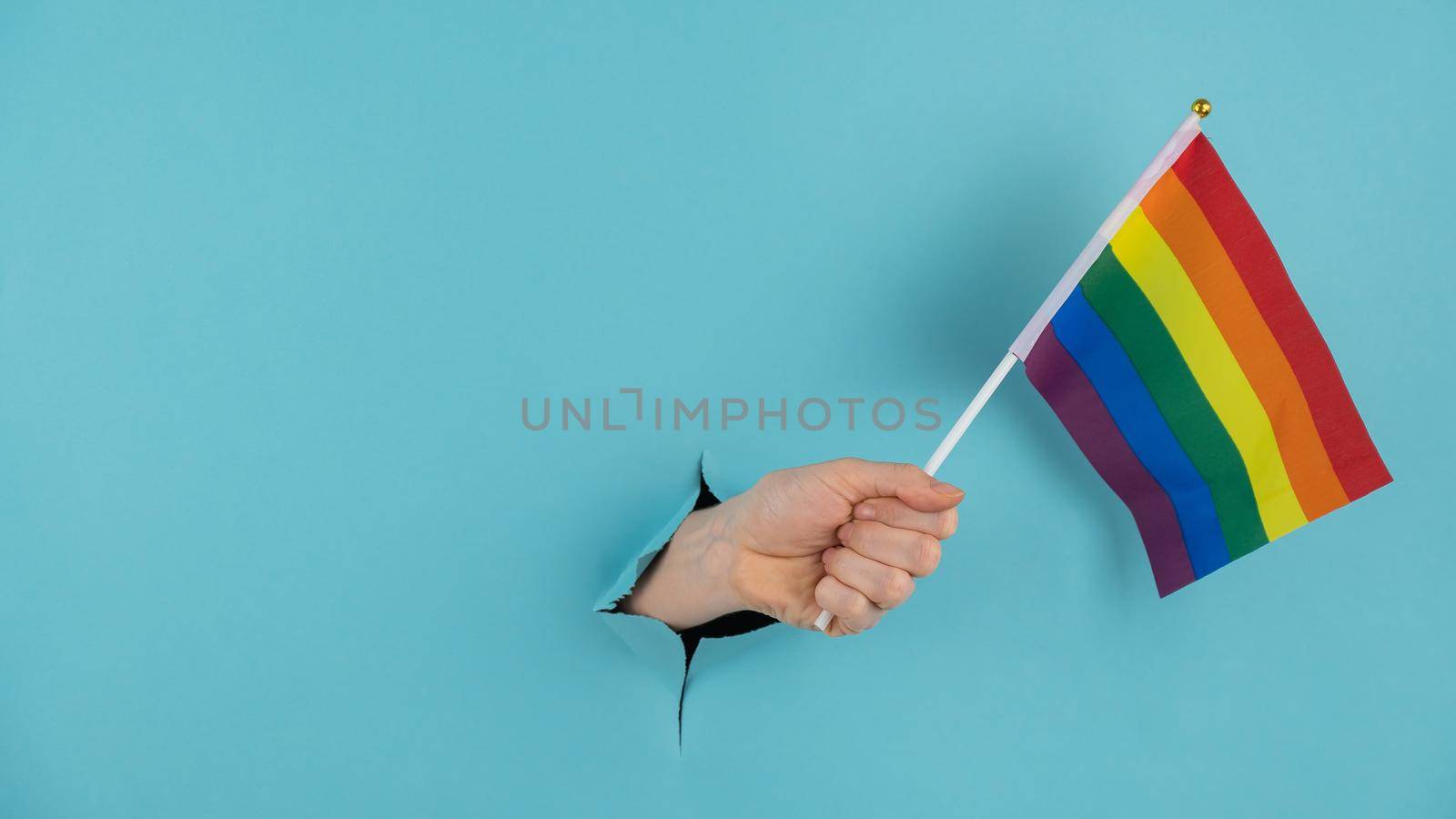 A woman's hand sticks out of a hole in a blue paper background and holds an lgbt flag