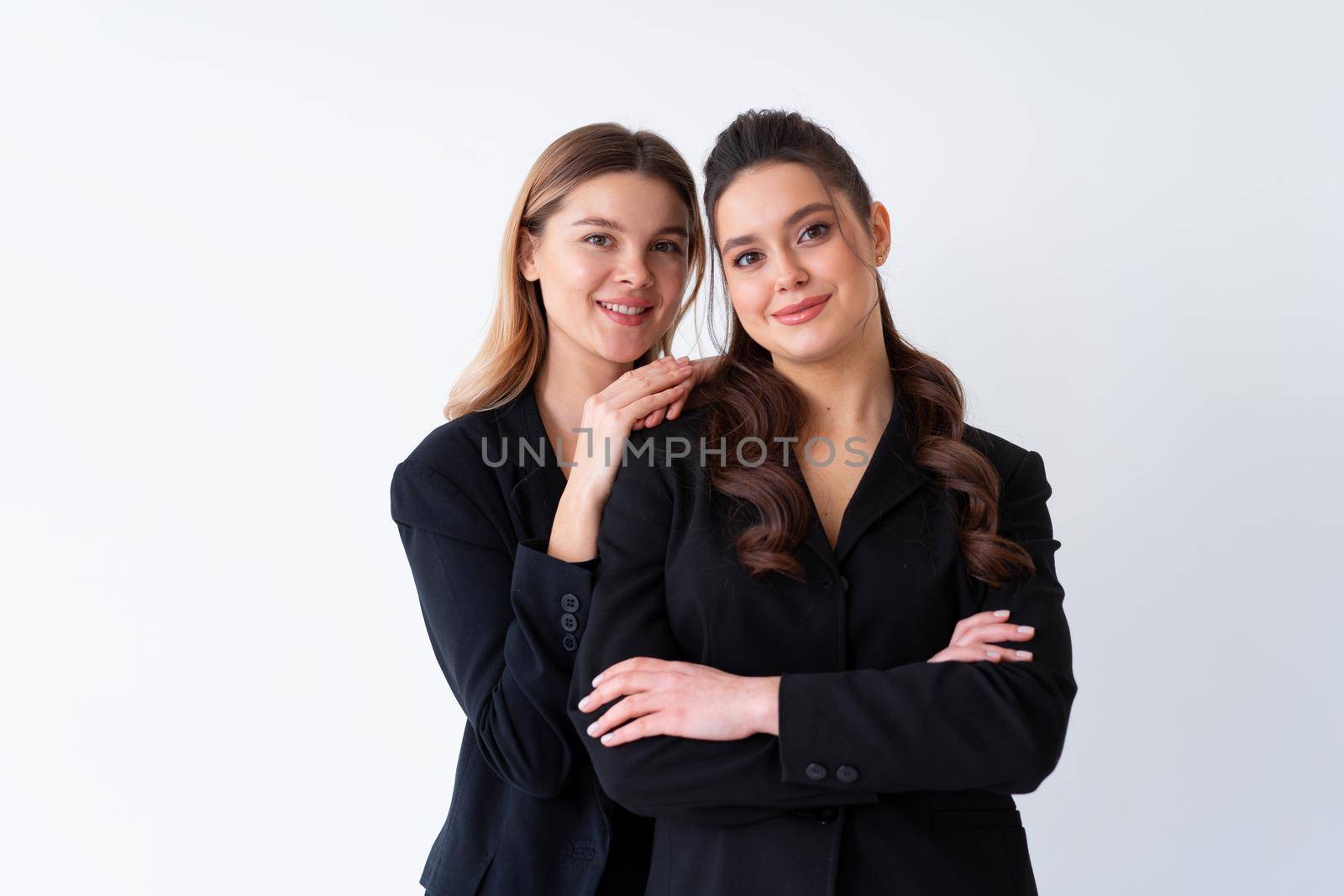 Concept of partnership in business. Two Young businesswoman dressed black formal suit standing studio isolated white background.Blonde woman put her hands on the shoulders of her friend. teeth Smile