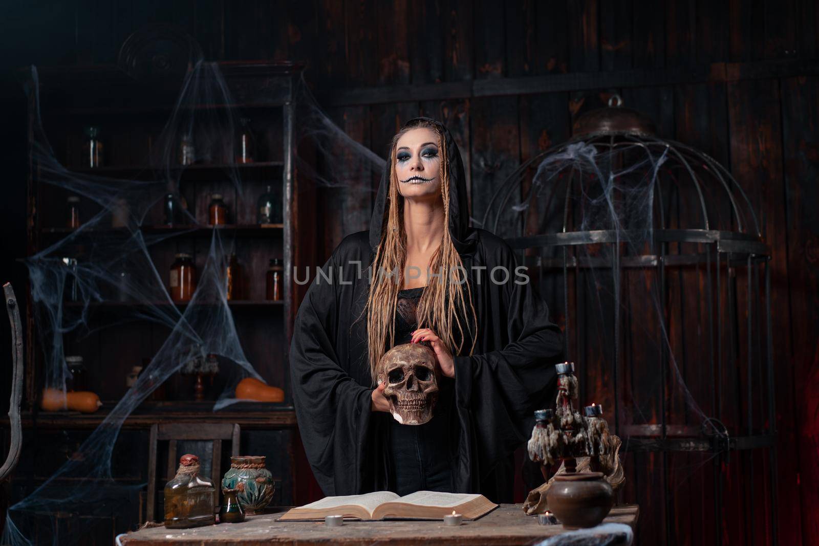 Halloween concept. Witch dressed black hood holding skull in hand standing dark dungeon room use magic book for conjuring magic spell. Female necromancer wizard gothic interior