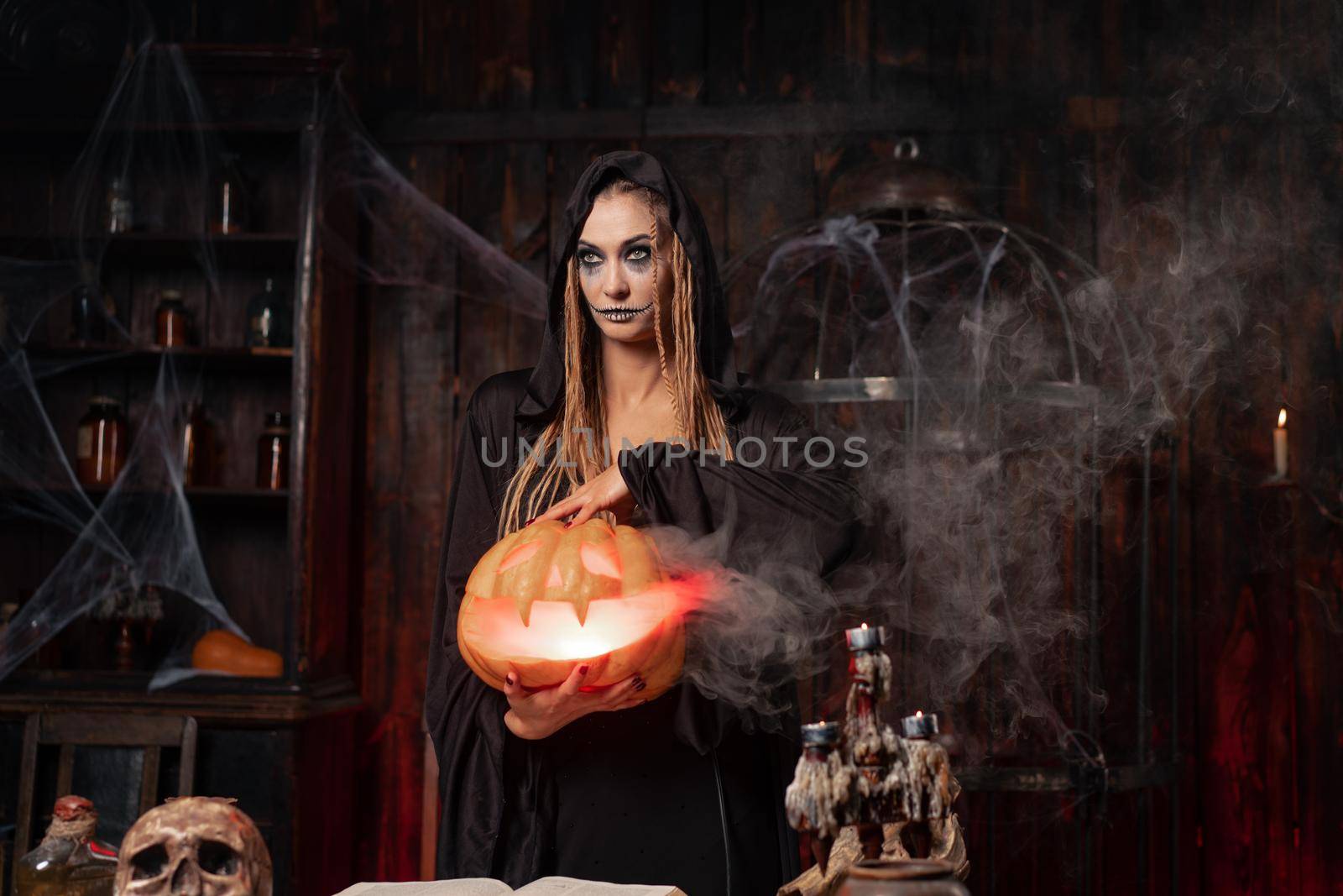 Halloween concept. Black witch holding Halloween pumpkin in hand with carved smily face in hand standing dark room. Female necromancer, dungeon place. Jack o lantern head. Wizard costume for party