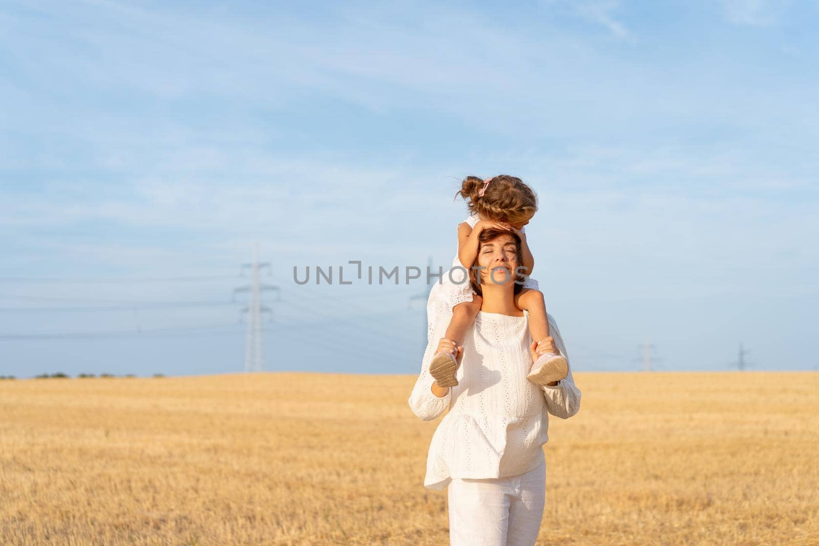 Little girl sitting mother shoulders hug her head. Little daughter piggyback mom standing wheat field blue sky background. Happy family concept. Love emotions. Tender. Dressed white dress. Together