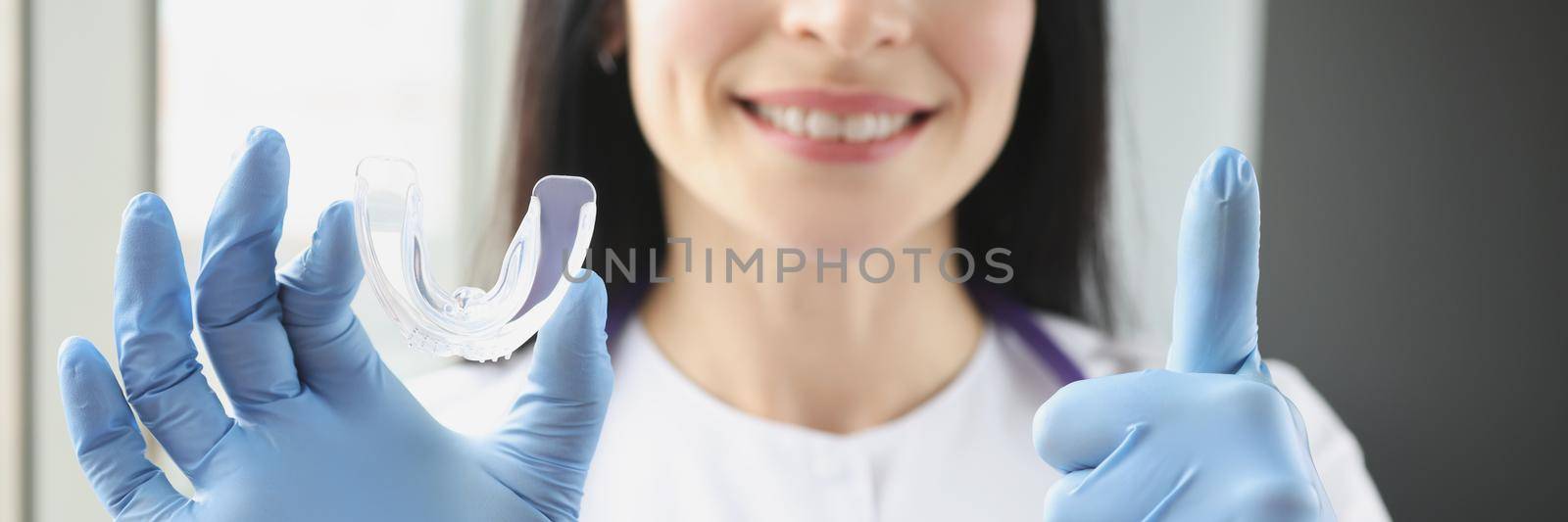 Portrait of stomatologist holding mouthguard for teeth and showing thumb up sign. Dentist at clinic office. Teeth whitening, dental treatment and dentistry concept