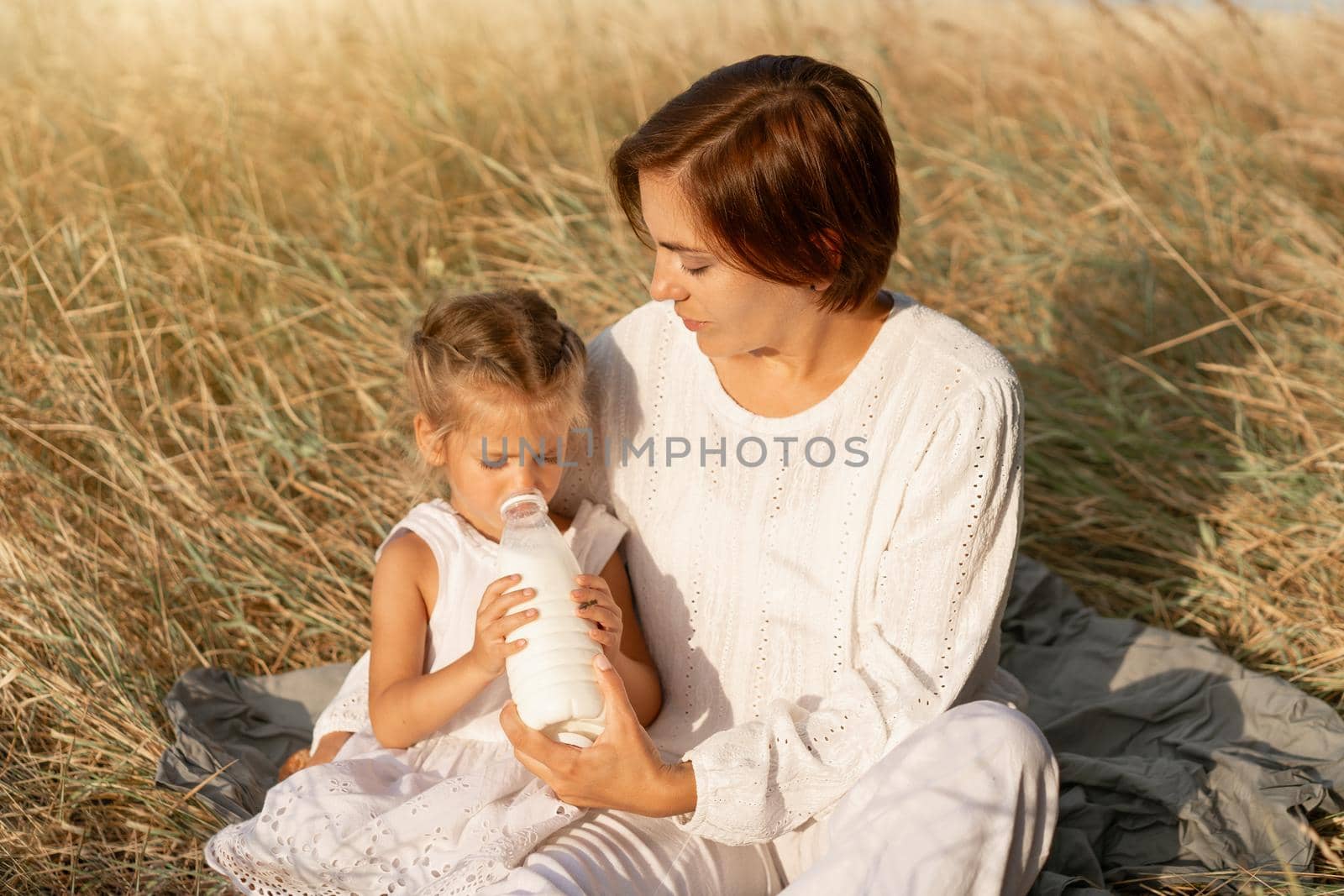 Little girl with her mother drink milk plastic bottle. Happy family, daughter with mom resting outdoor drink healthy diary product. Countryside rural scene. Caucasian female on nature