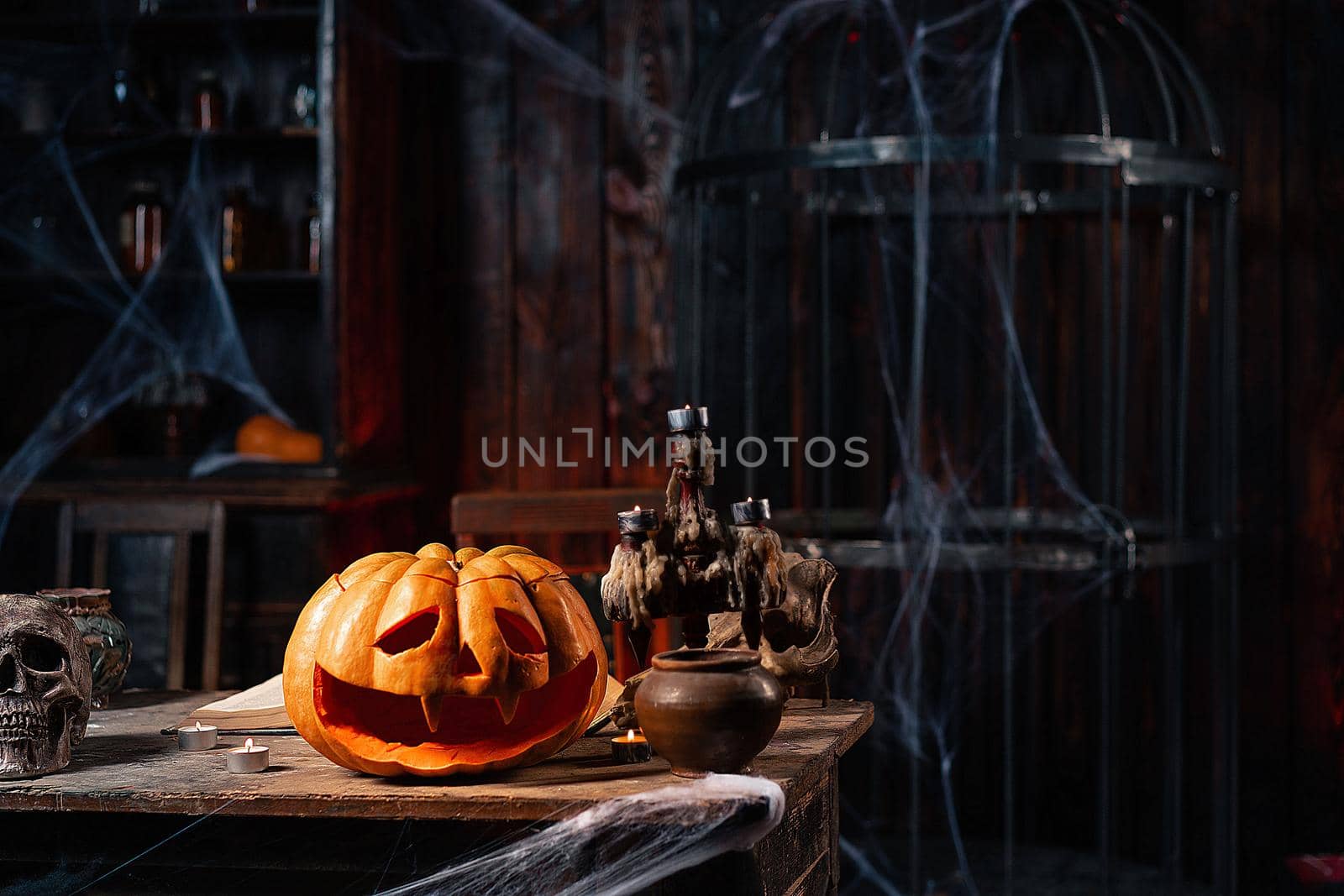 Halloween. Scary Halloween pumpkin with carved face on table in dark room with candles, spider web, and cage on background. Copy space for text. Spooky atmosphere. Mysterious room in dark dungeon