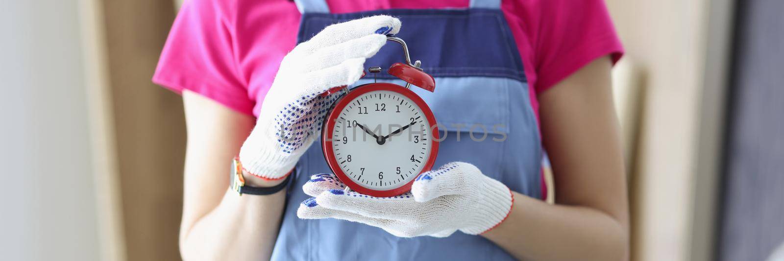 Close-up of woman worker in uniform holding red clock reminder to finish work on time. Foreman at construction site control progress. Renovation concept