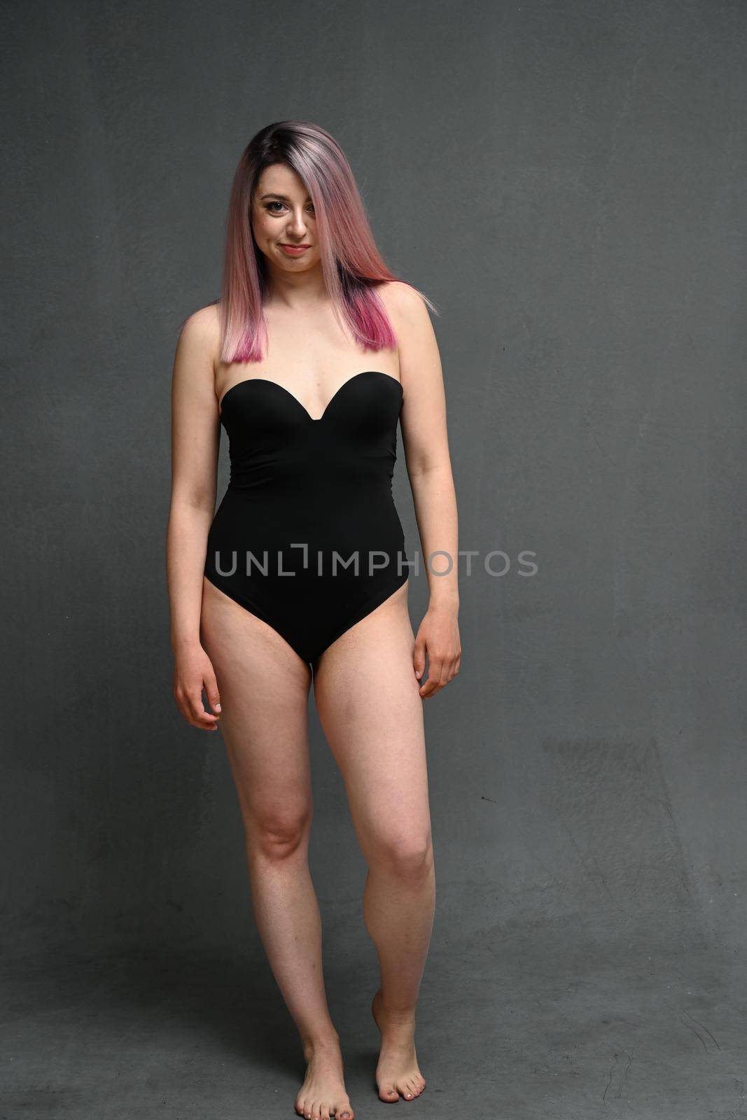 Horizontal photo of a caucasian woman with dyed hair in full growth in a black bodysuit with a smile looks at the camera on a gray background in the studio by chichaevstudio