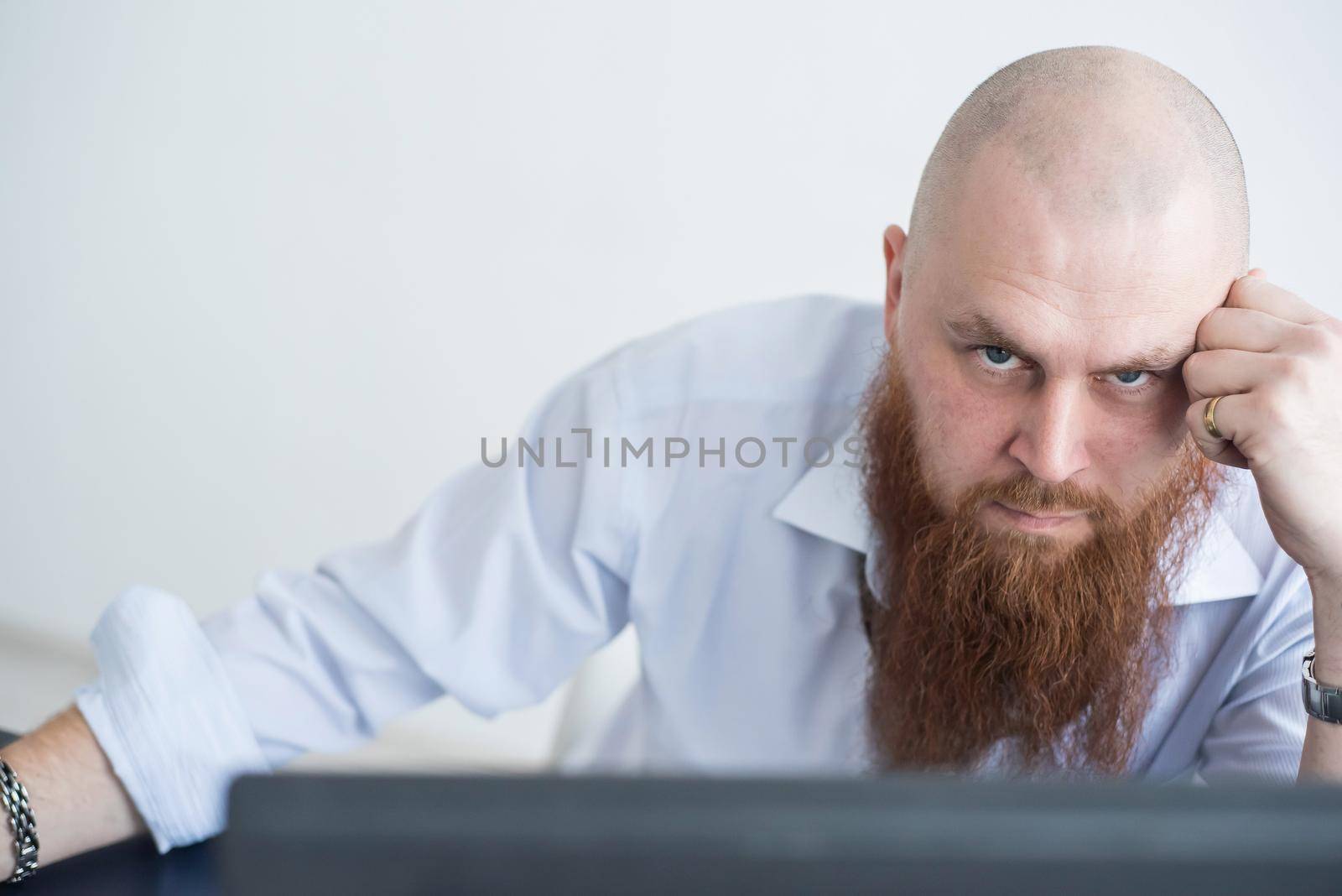 A focused bald man with a red beard stares intently at the camera. Male manager in a white shirt is angry at work