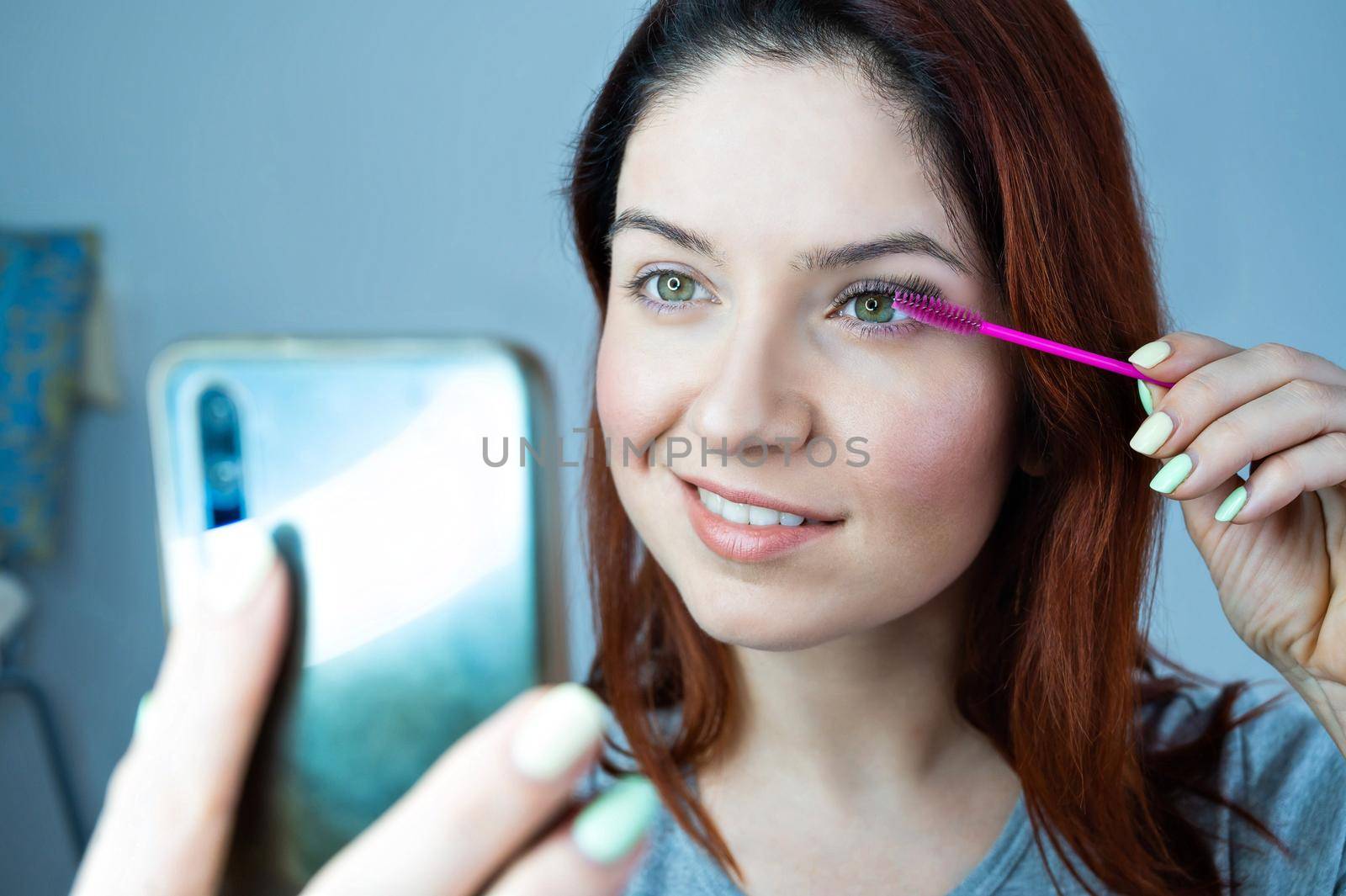 Beautiful redhead woman paints eyelashes and takes a selfie. The girl puts mascara and looks at the phone instead of a mirror. by mrwed54