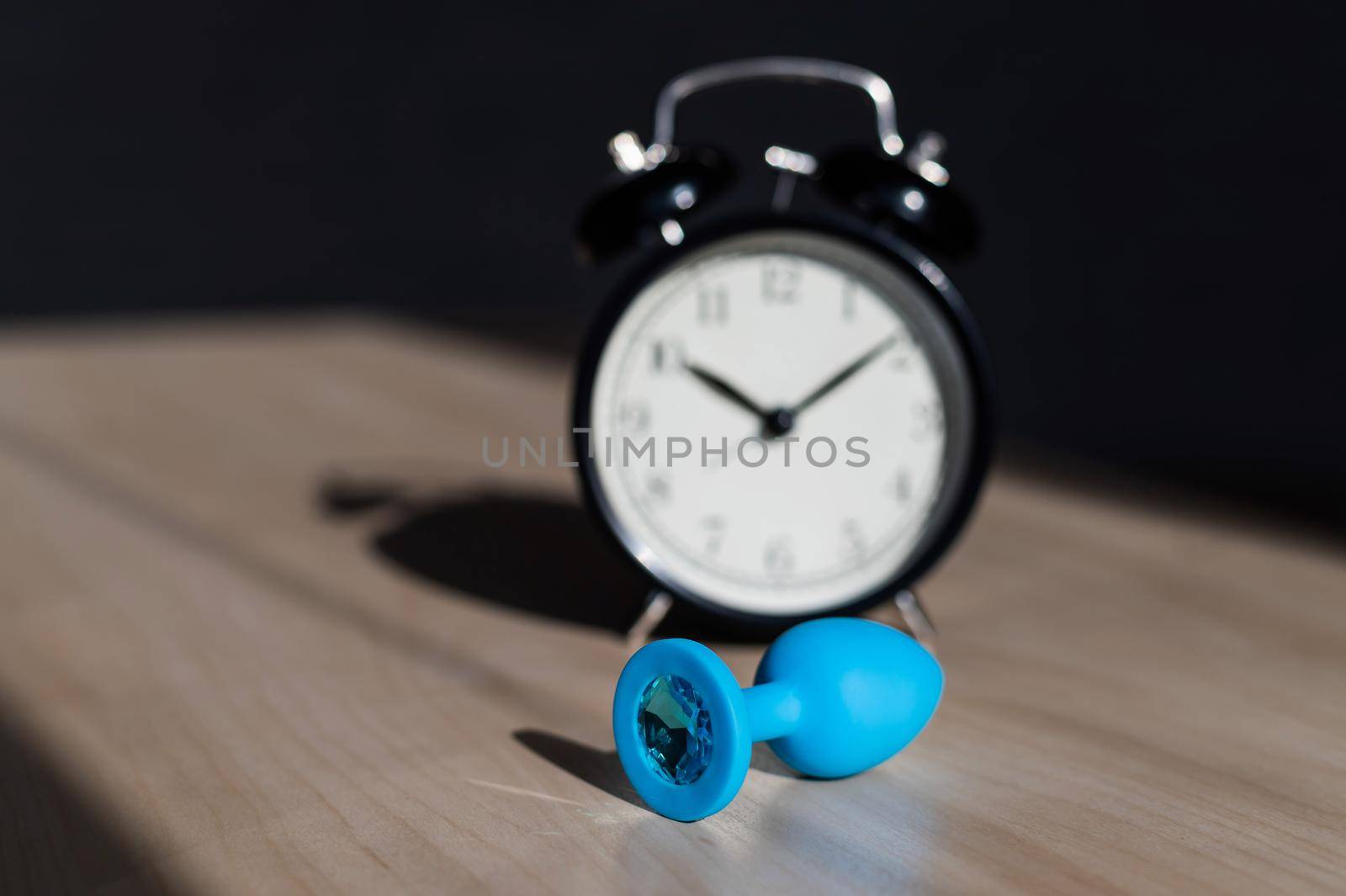 Blue latex anal plug with crystals and an alarm clock on a wooden table. The time of carnal pleasures